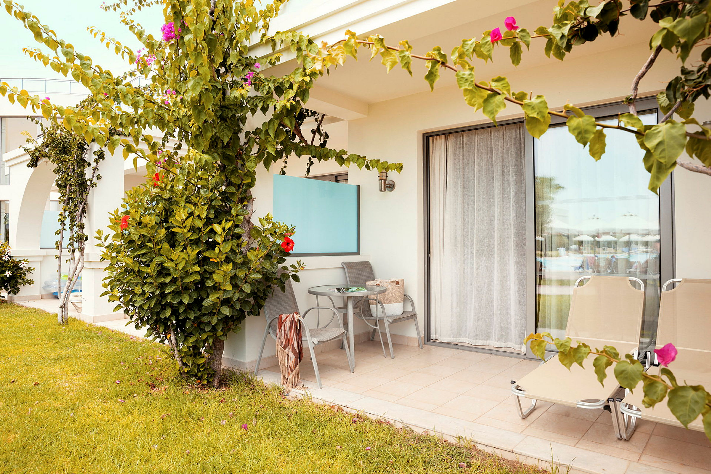 Terrasse med bord, stoler og solsenger. Foran er det gressplen og blomstrende planter. (AI-generert tekst)