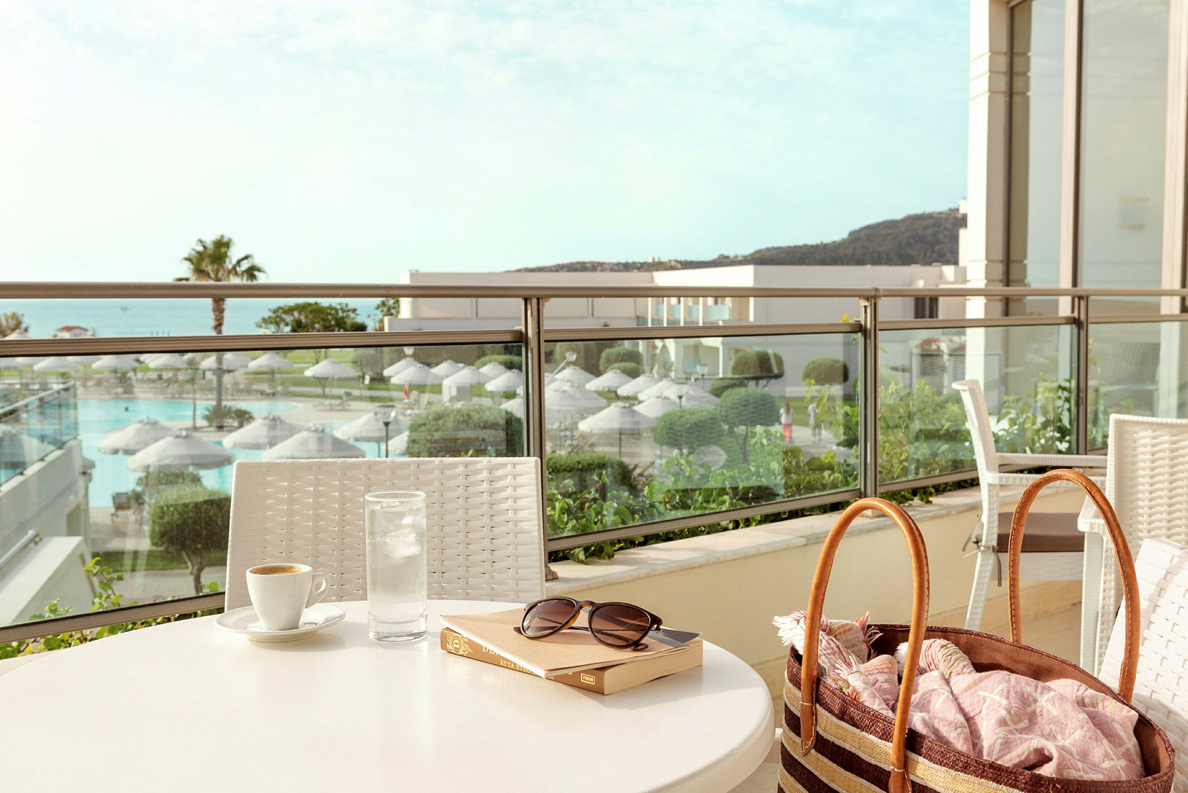 Et bord med en kopp kaffe, et glass vann og en bok plassert på en terrasse med utsikt over et bassengområde og beplantning. (AI-generert tekst)