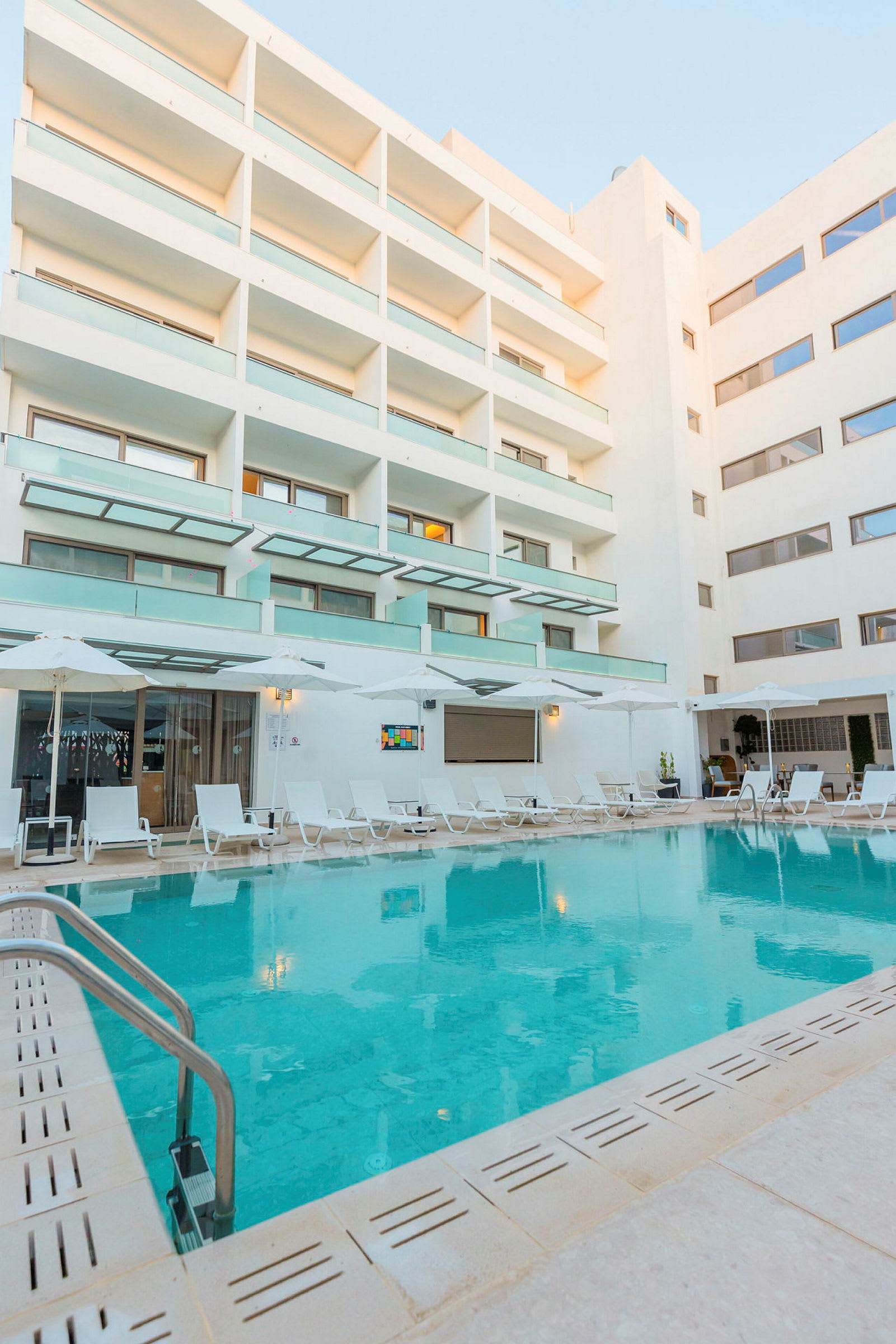 Et hotellbasseng omgitt av solsenger og en moderne hotellbygning. (AI-generert tekst)