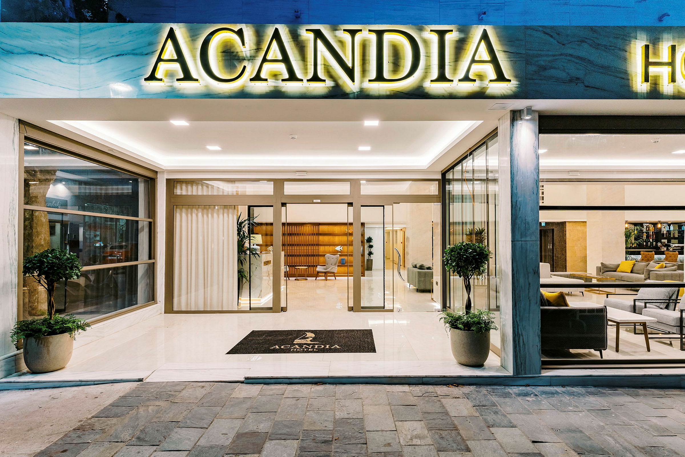 Inngangen til Hotel Acandia med glassdører og et skilt med hotellets navn. (AI-generert tekst)