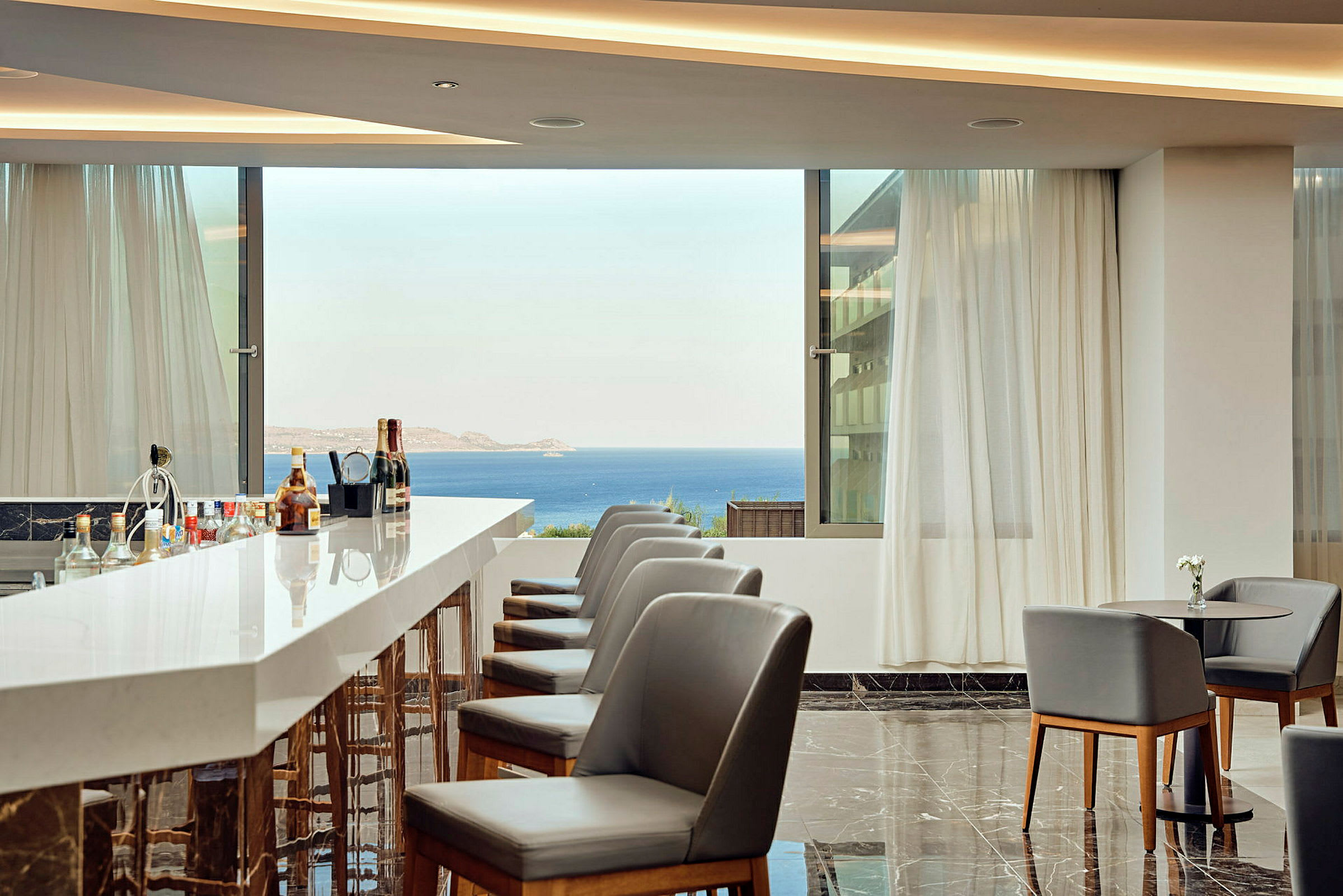 En hotellbar med moderne stoler, marmorgulv og utsikt over havet fra store vinduer. (AI-generert tekst)