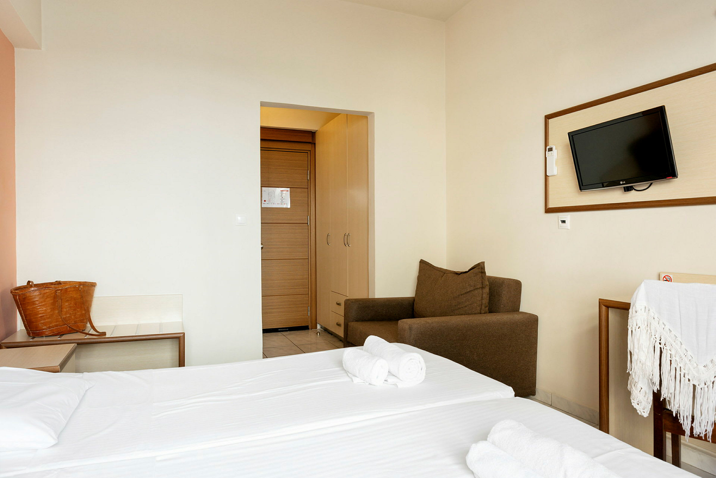 Hotellrom med dobbeltseng, brun lenestol, vegghengt TV og dør med garderobe. (AI-generert tekst)