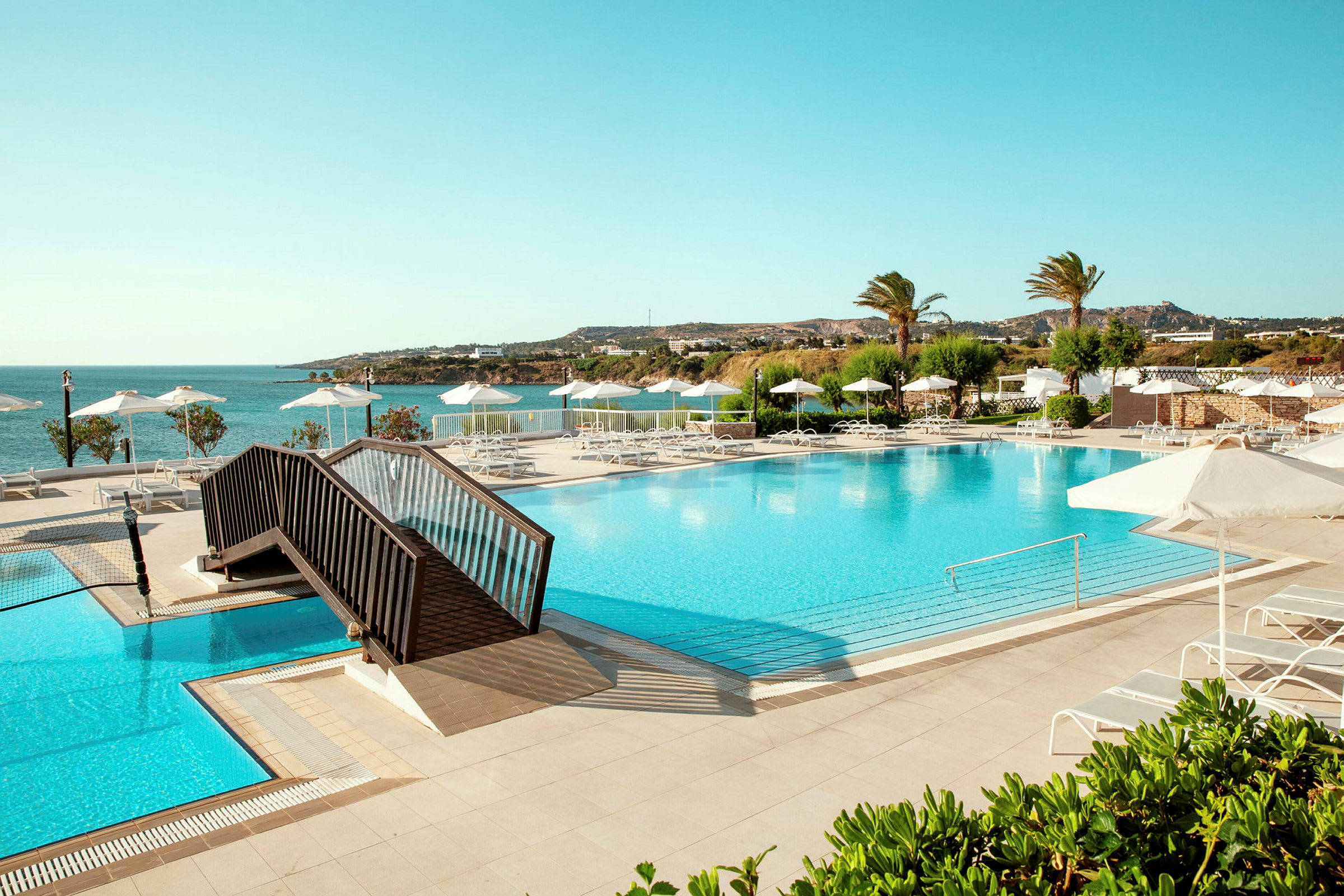 Stor basseng med solsenger og parasoller, utsikt mot sjøen og vegetasjon. (AI-generert tekst)