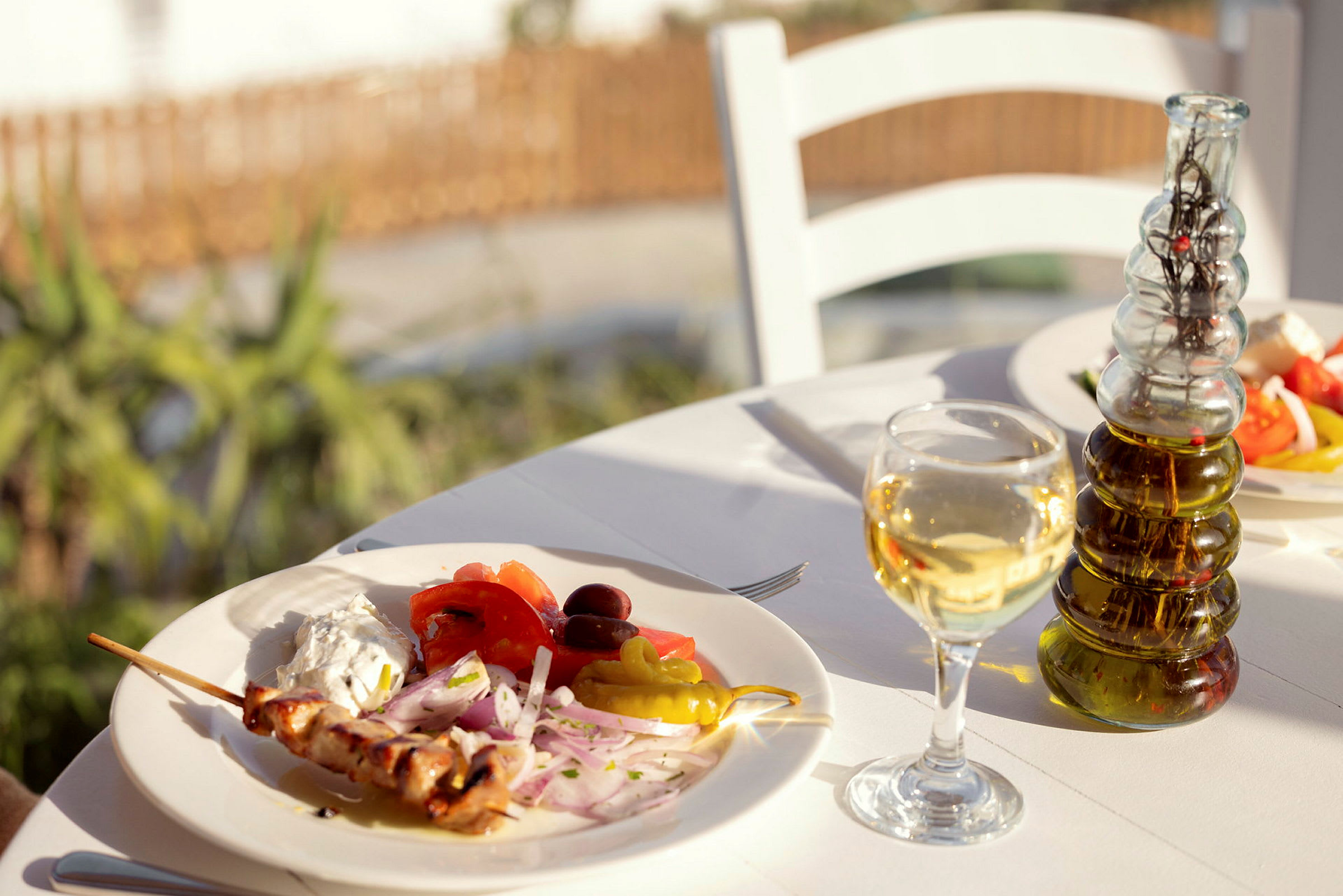 En tallerken med grillspyd, grønnsaker og saus på et bord utendørs, med et glass hvitvin og en flaske olivenolje. (AI-generert tekst)