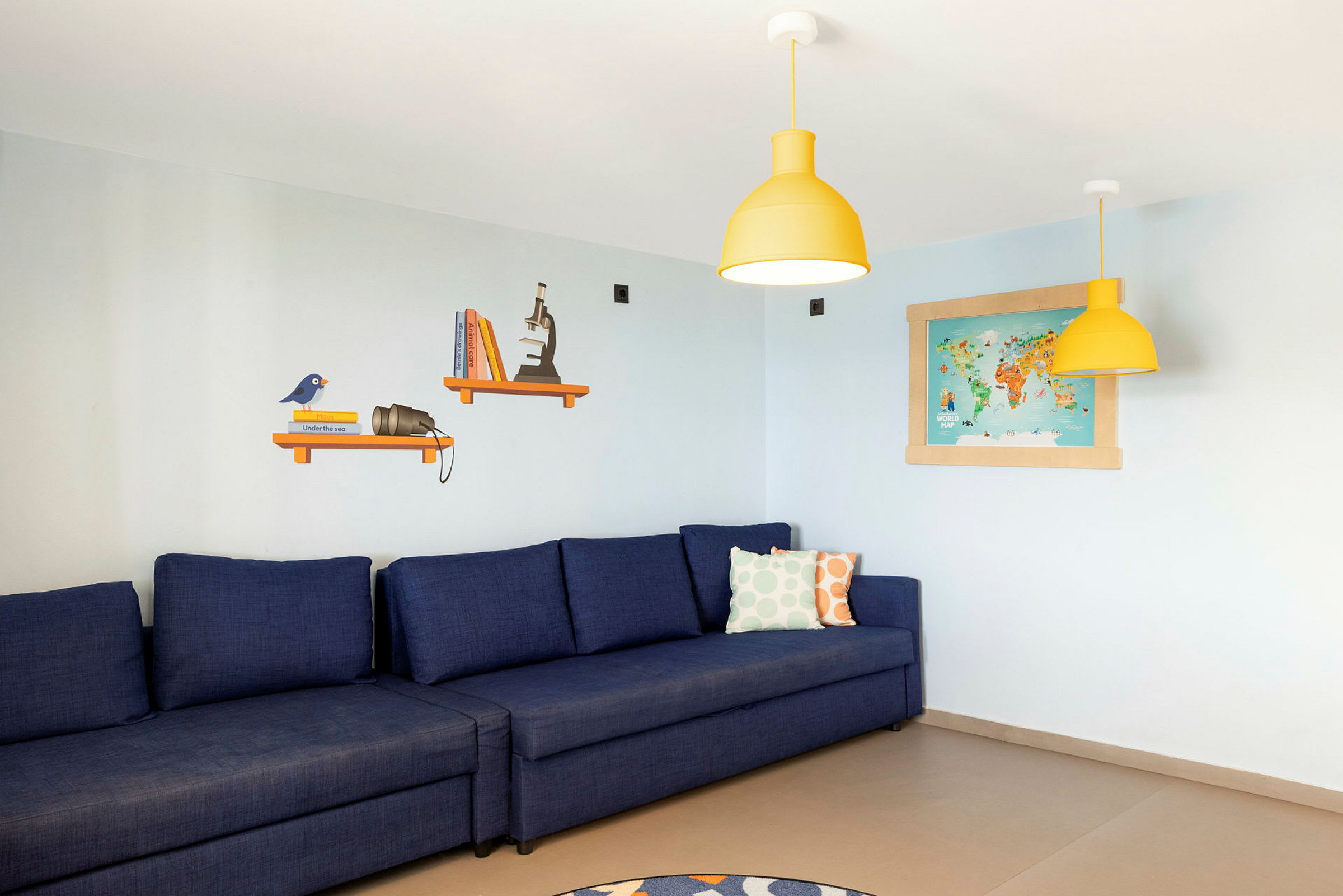 En blå sofa med fargede puter står mot en lys vegg med hyller, lamper og et verdenskart. (AI-generert tekst)