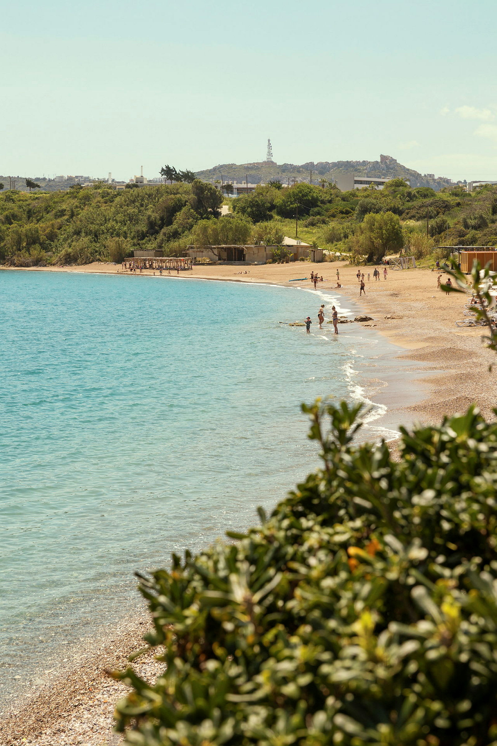 Utsikt over en strand med turkis vann og badende mennesker, omgitt av grøntområder. (AI-generert tekst)