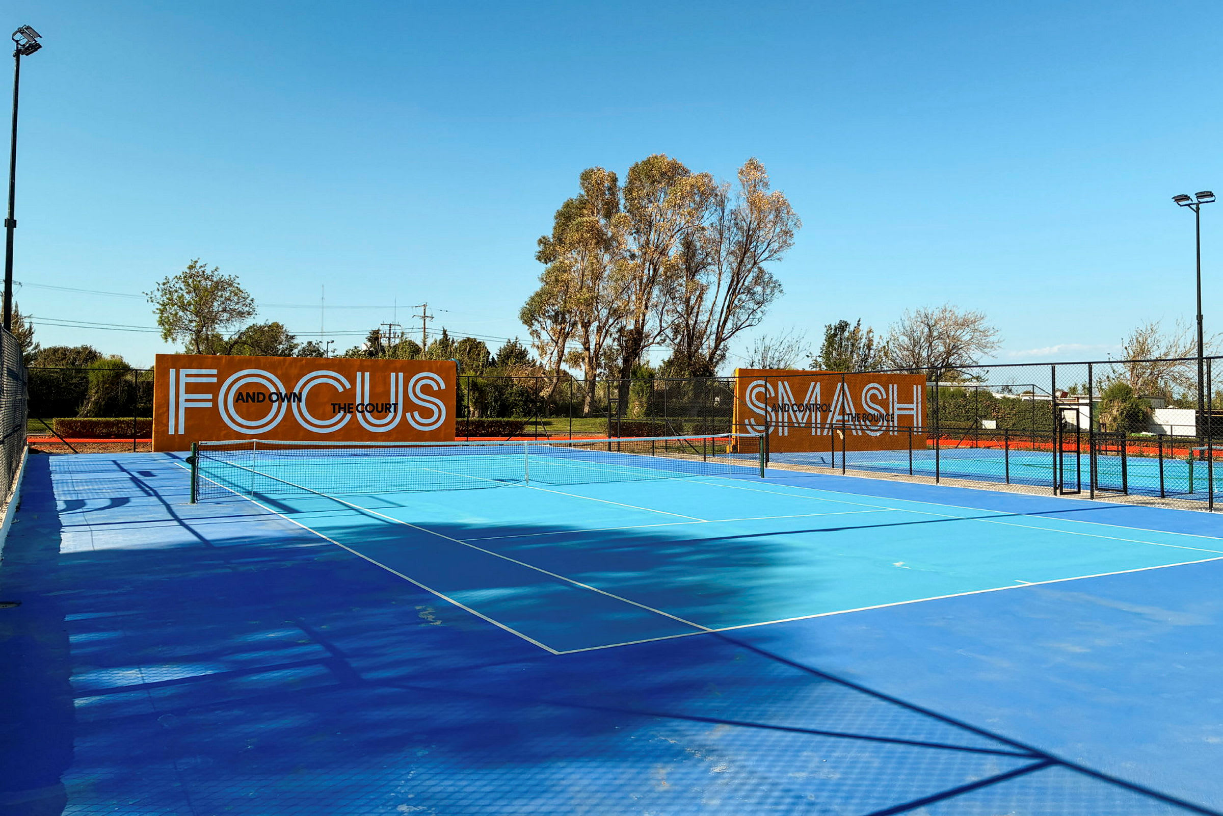 Blå tennisbane utendørs med trær i bakgrunnen og skilt som viser teksten "Focus" og "Smash". (AI-generert tekst)