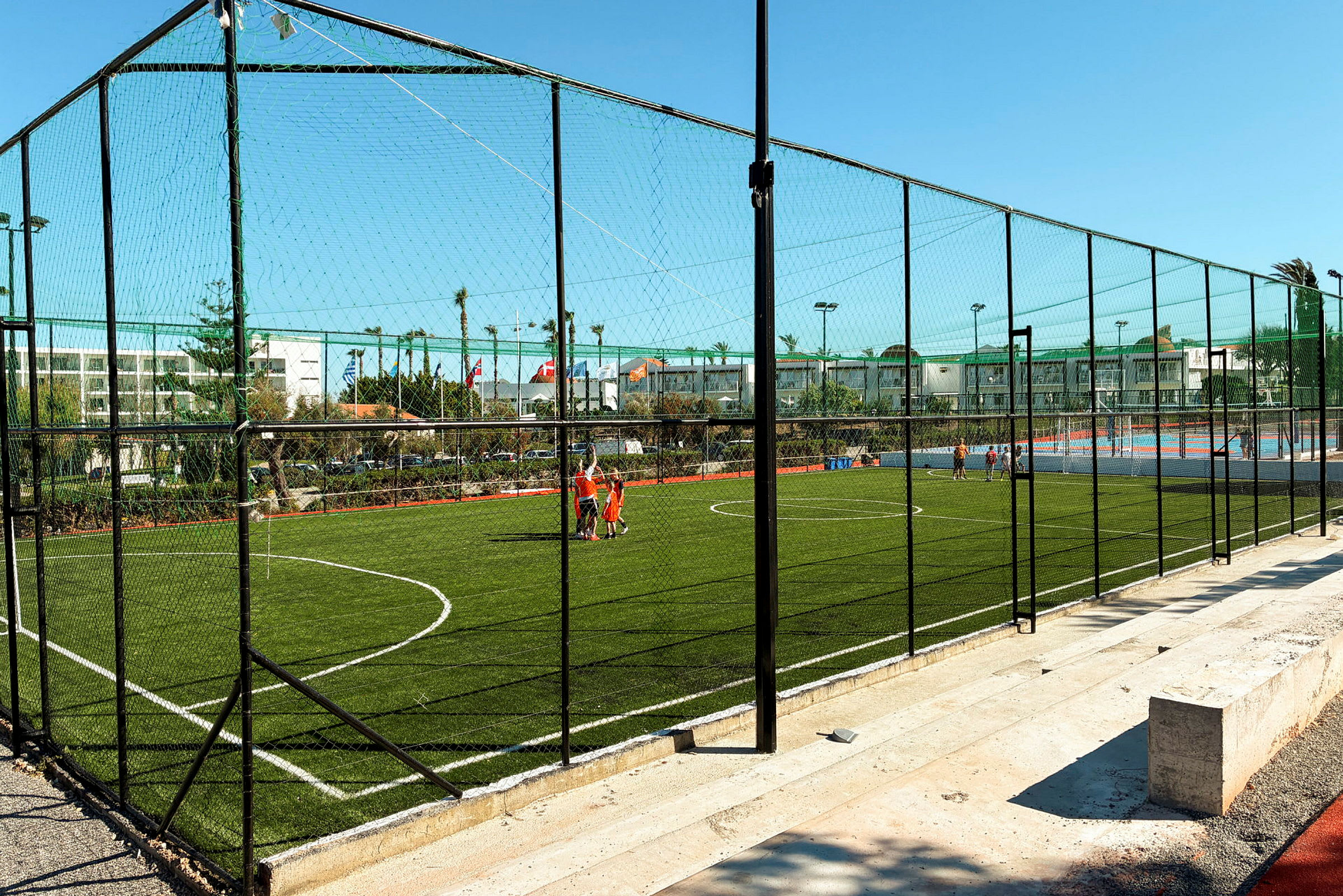 En fotballbane med kunstgress omgitt av et beskyttelsesnett og gjerde, hvor noen mennesker spiller fotball. (AI-generert tekst)