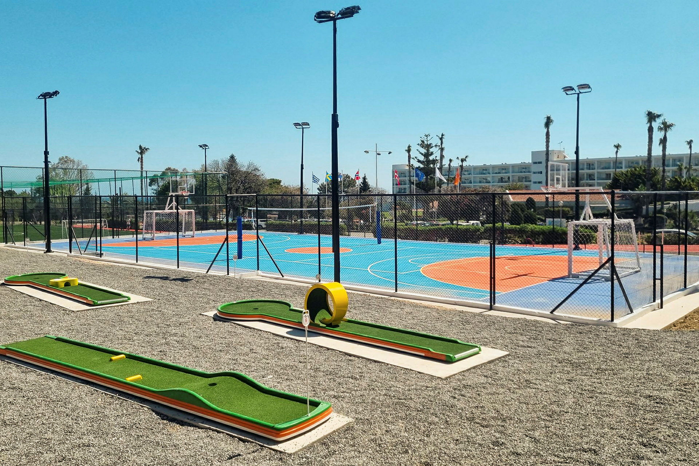 Et bilde av et utendørsmiljø med en minigolfbane i forgrunnen og en fargerik sportsbane med oransje og blå overflate omgitt av gjerde i bakgrunnen. (AI-generert tekst)