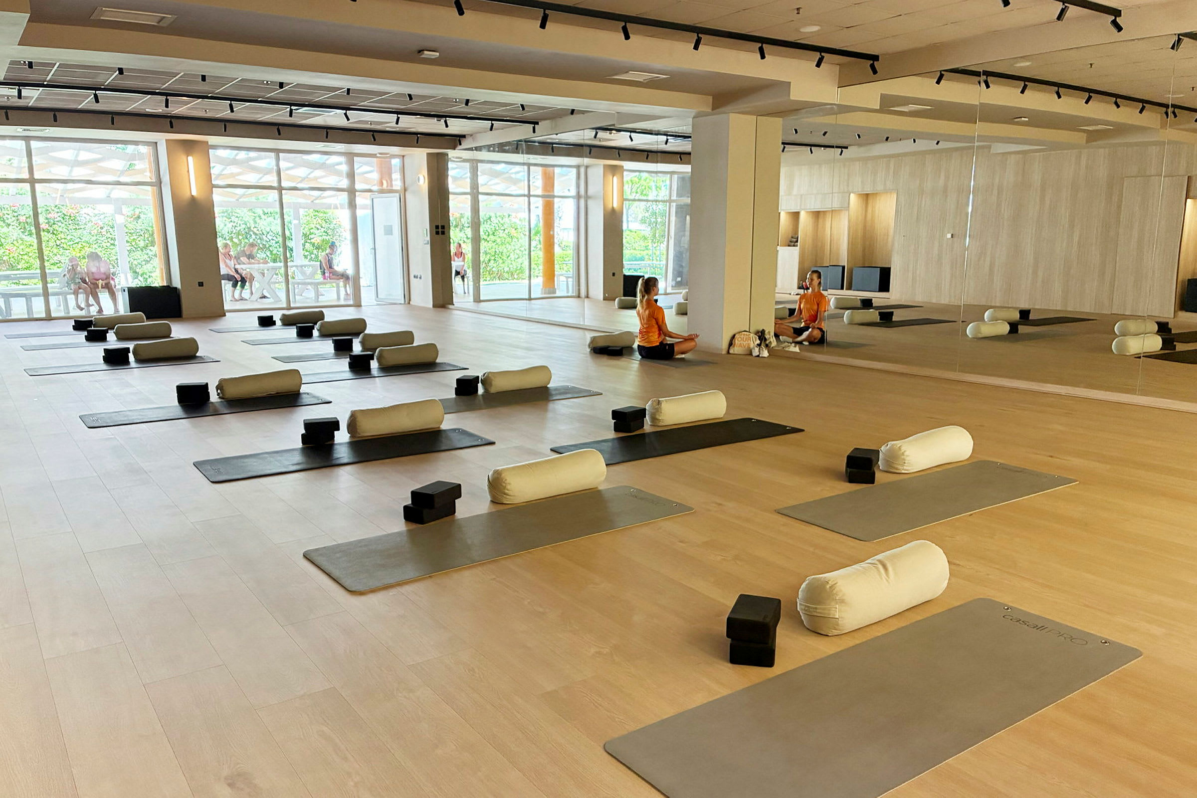 Et treningsrom med yogamatter og tilbehør som puter og blokker lagt ut. To personer har en samtale ved speilet i bakgrunnen. (AI-generert tekst)