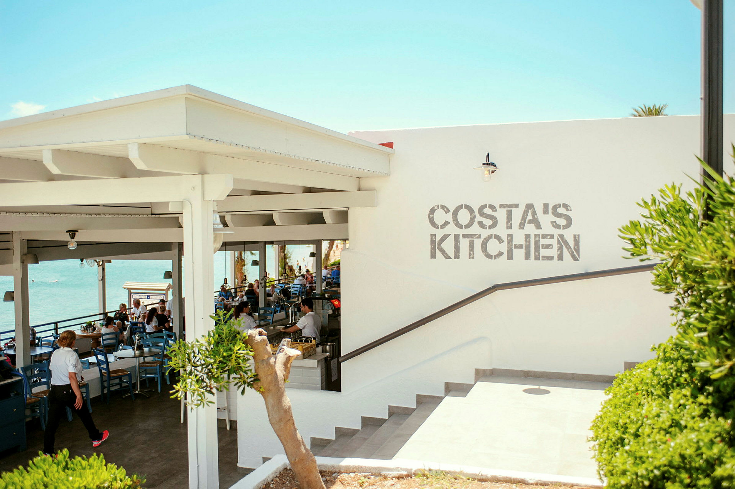 En utendørs restaurant med utsikt over havet og teksten 'Costa's Kitchen' på veggen. (AI-generert tekst)