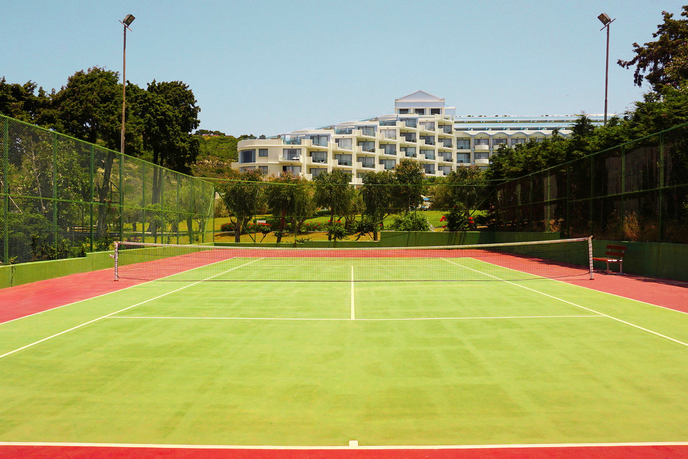 En grønn tennisbane med et nett i midten og et hotell i bakgrunnen omgitt av trær. (AI-generert tekst)