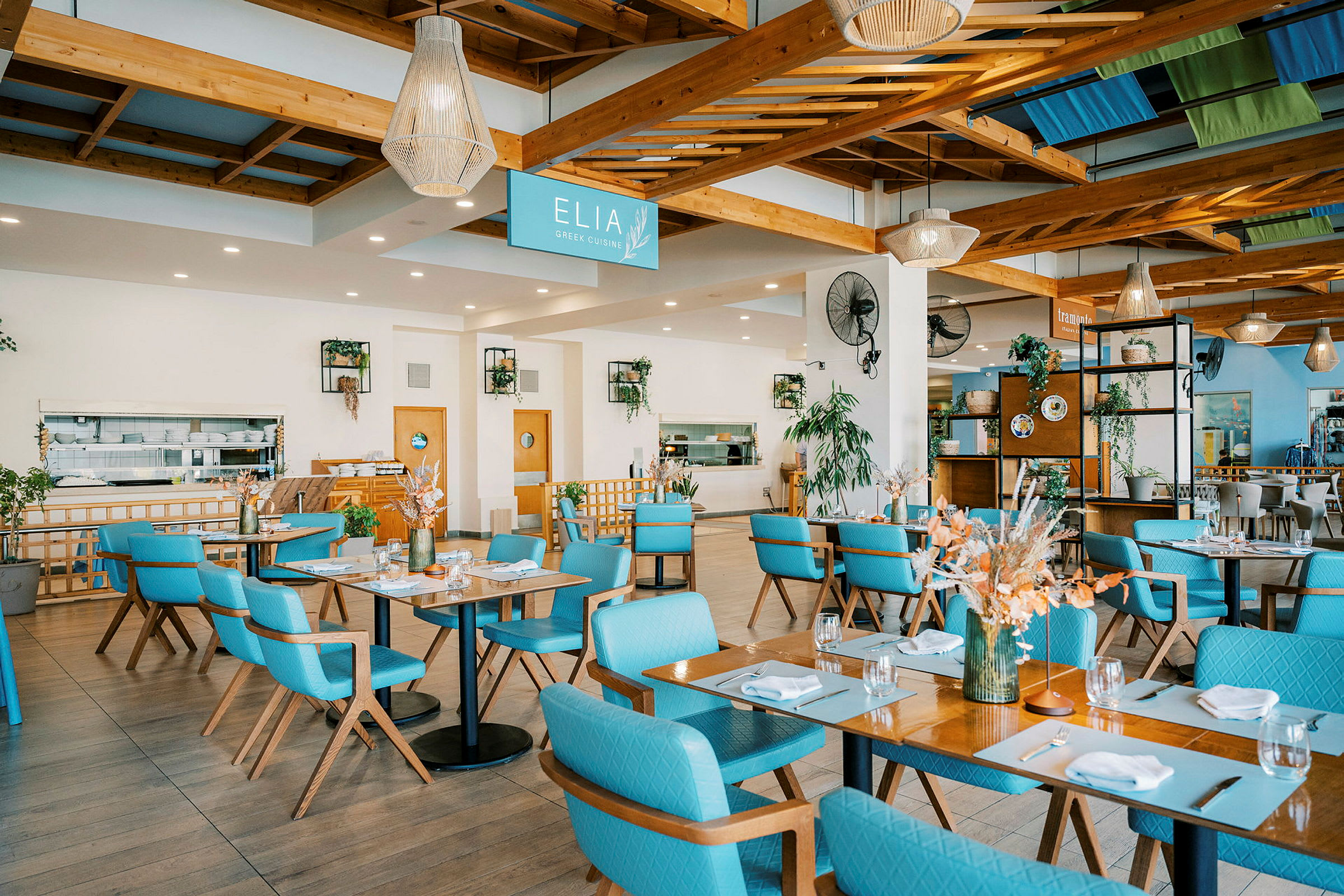 En lys og romslig restaurantmiljø med bord og blå stoler. Dekorasjoner og planter er en del av innredningen. (AI-generert tekst)