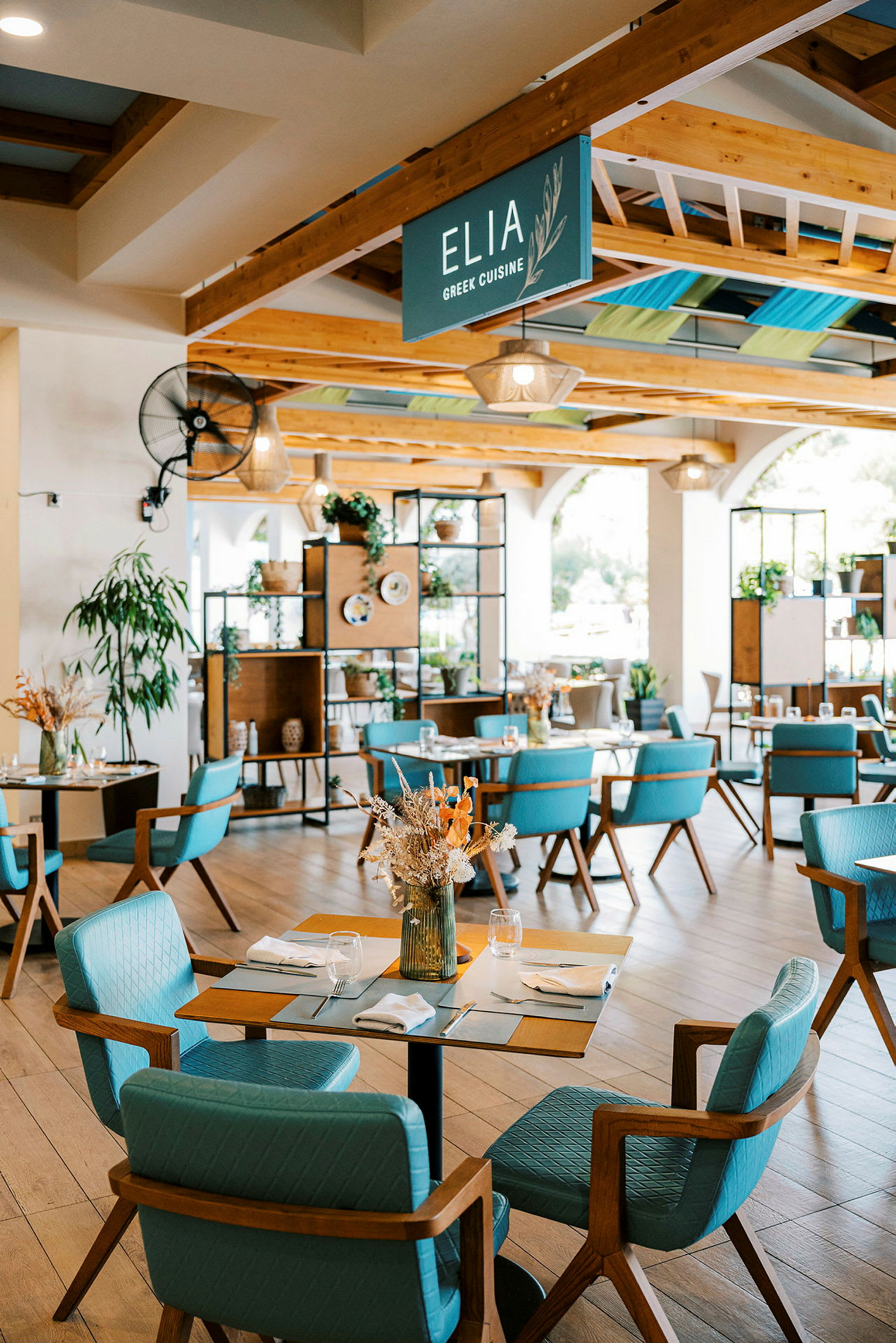 En lys og moderne restaurant med tredetaljer og blå stoler. Dekorert med planter og tørkede blomsteroppsatser. (AI-generert tekst)
