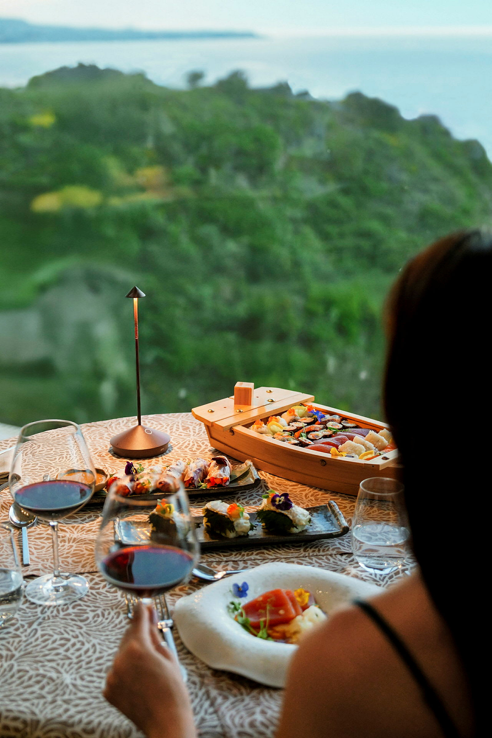 Et vakkert dekket måltid med sushi og vin står på et bord med utsikt over et grønt landskap og havet. (AI-generert tekst)