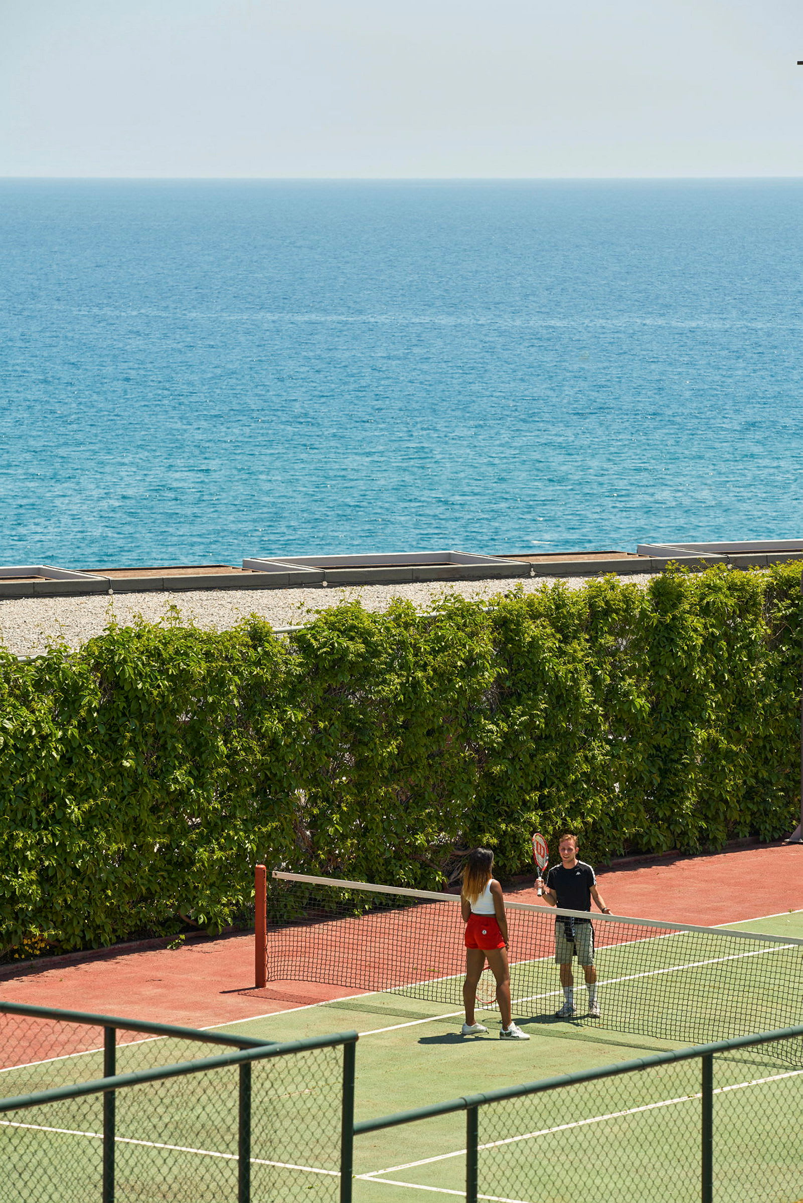To personer spiller tennis på en tennisbane nær sjøen. (AI-generert tekst)