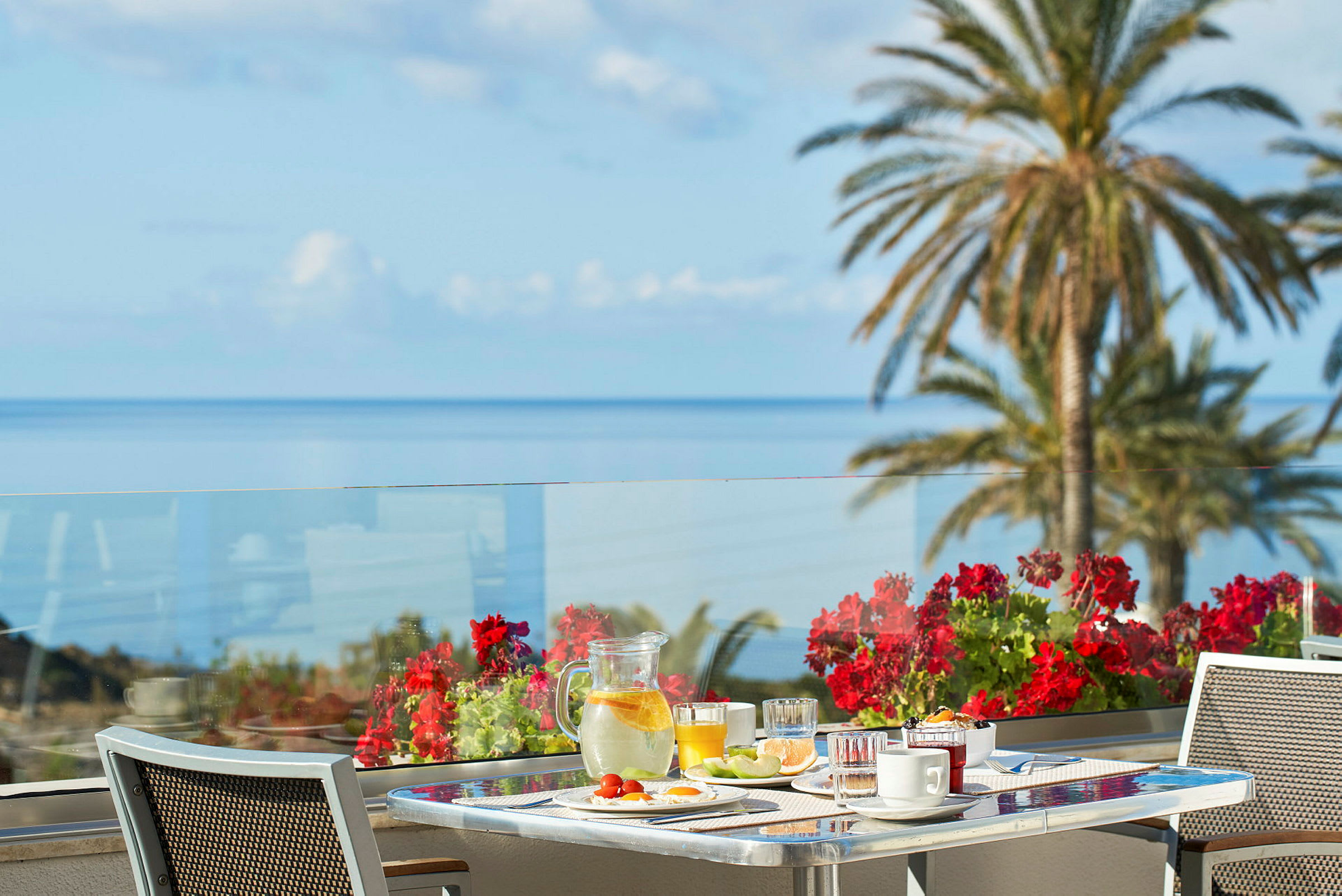 En frokostservering på en terrasse med havutsikt og røde blomster i fokus. (AI-generert tekst)