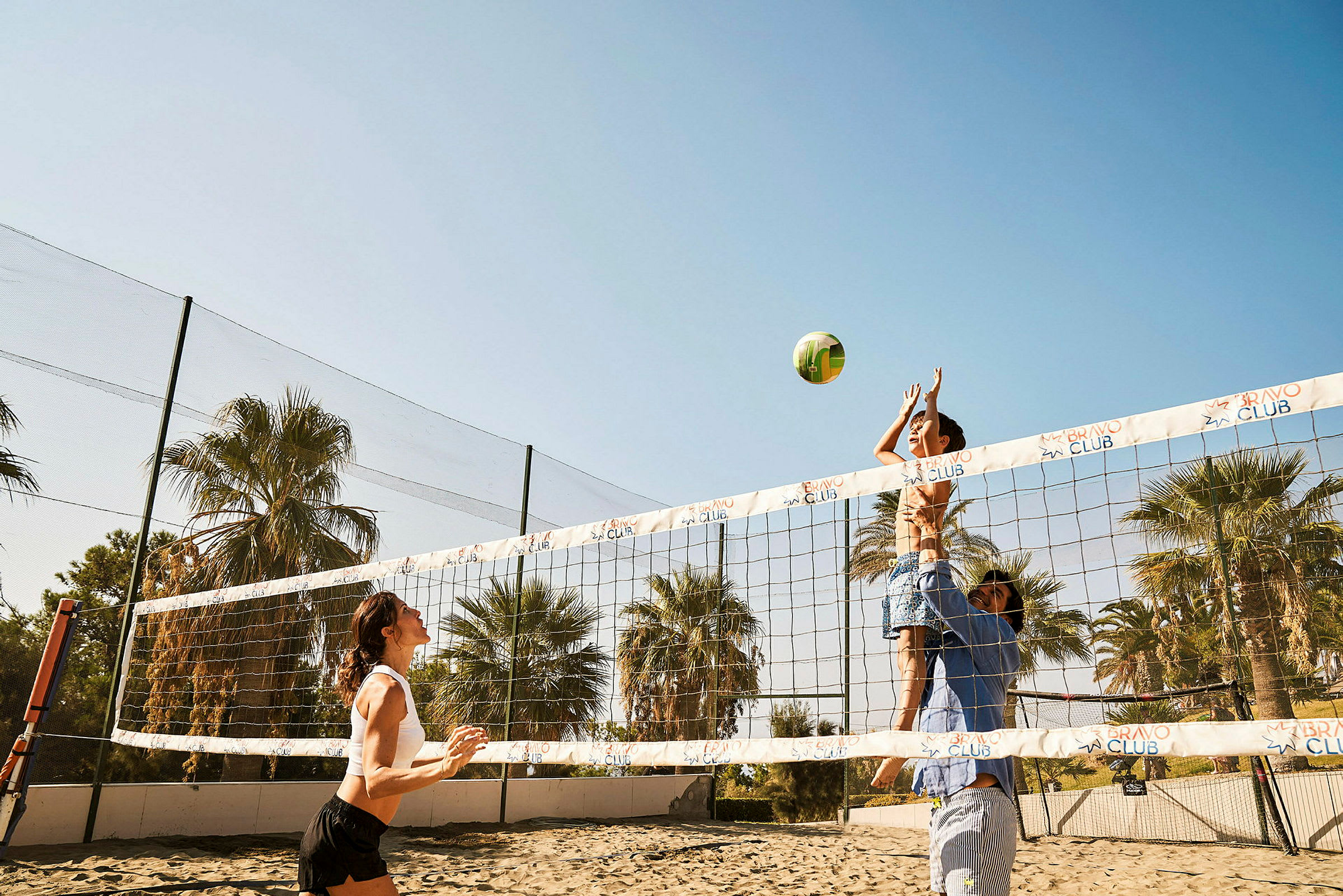 Personer spiller volleyball på en sandbane med palmer i bakgrunnen. (AI-generert tekst)