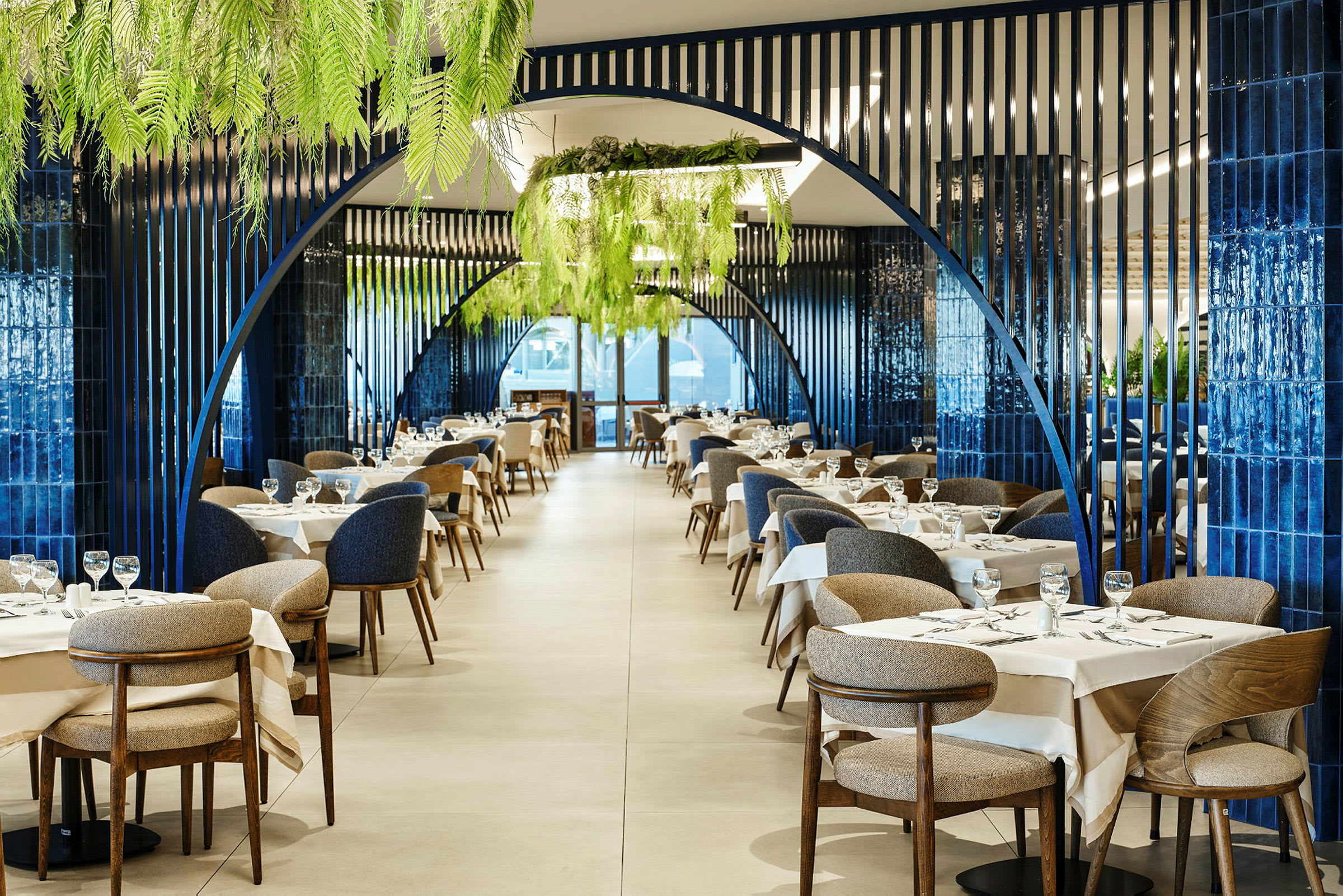 En elegant innredet restaurant med blå buestrukturer, hvite duker og grønne planter i taket. (AI-generert tekst)