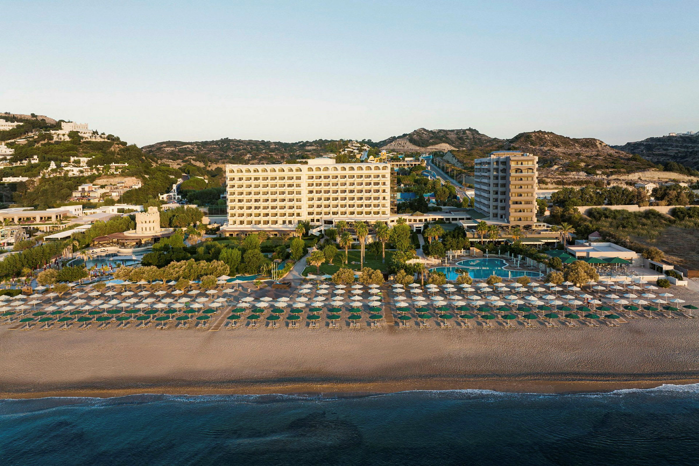 Et fleretasjes hotell bak en sandstrand fylt med parasoller, omgitt av trær og bassenger. (AI-generert tekst)