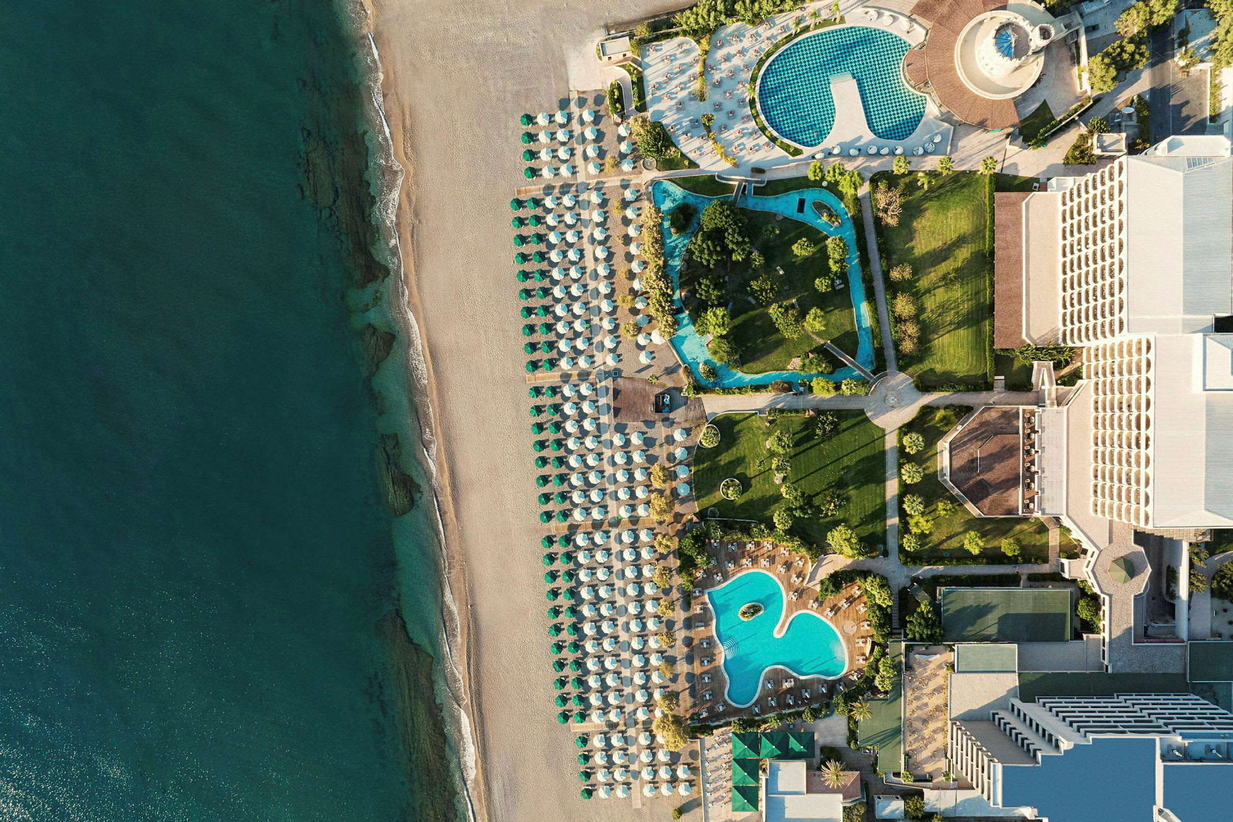 Luftfoto som viser en strand med parasoller, bassengområder og hotellbygginger. (AI-generert tekst)