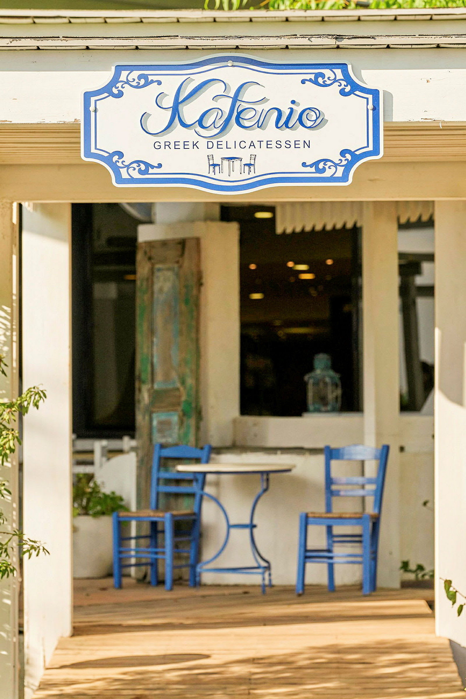 To blå stoler og et rundt bord står på en utendørsterrasse foran en bygning. Et skilt over døren lyder "Kafenio Greek Delicatessen". (AI-generert tekst)