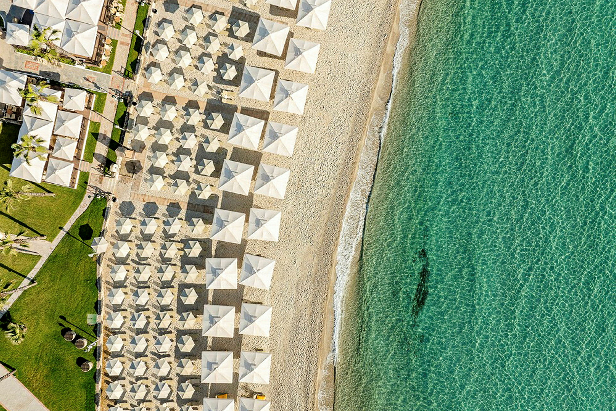 Flyfoto av en strand med solparasoller i rekker og klart turkis vann. (AI-generert tekst)