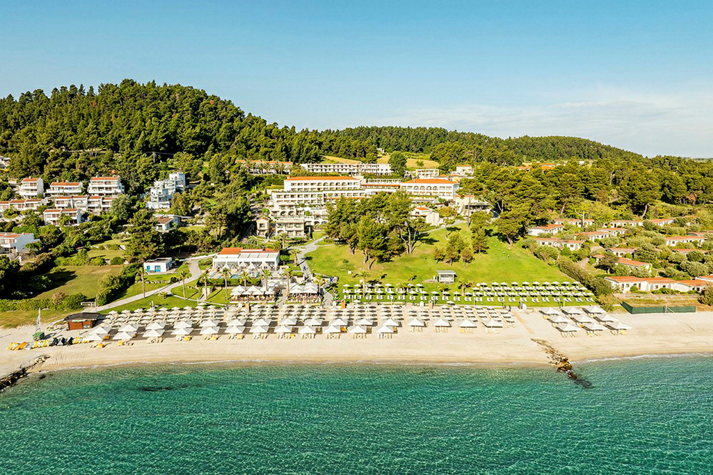 Luftfoto av en ferieresort ved stranden med solsenger og parasoller samt omkringliggende grønne skogkledde åser. (AI-generert tekst)