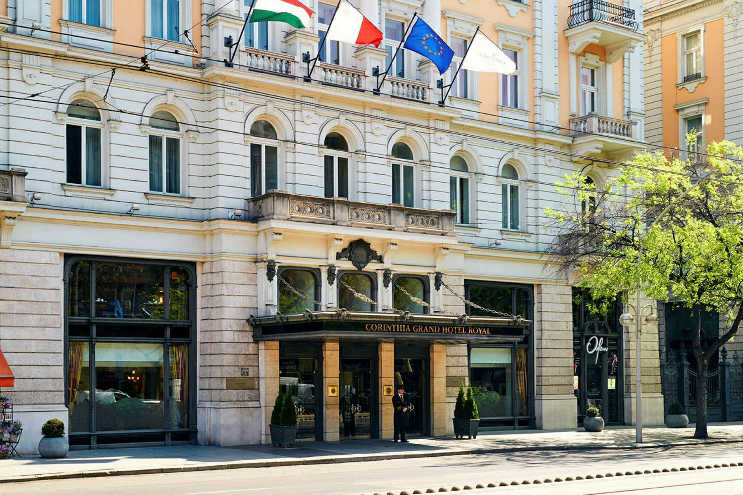 Forsiden av Corinthia Grand Hotel Royal med glassinngang og flagg over. (AI-generert tekst)