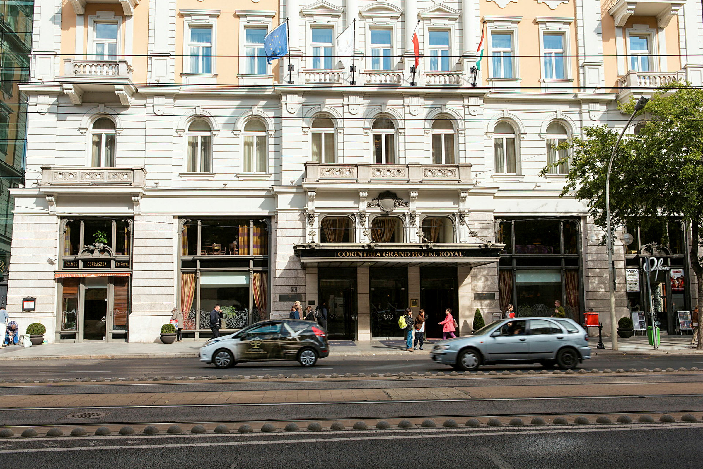 Fasade av et hotell med flere etasjer, innvendig synlige vinduer og inngangsområdet med mennesker og passerende biler. (AI-generert tekst)