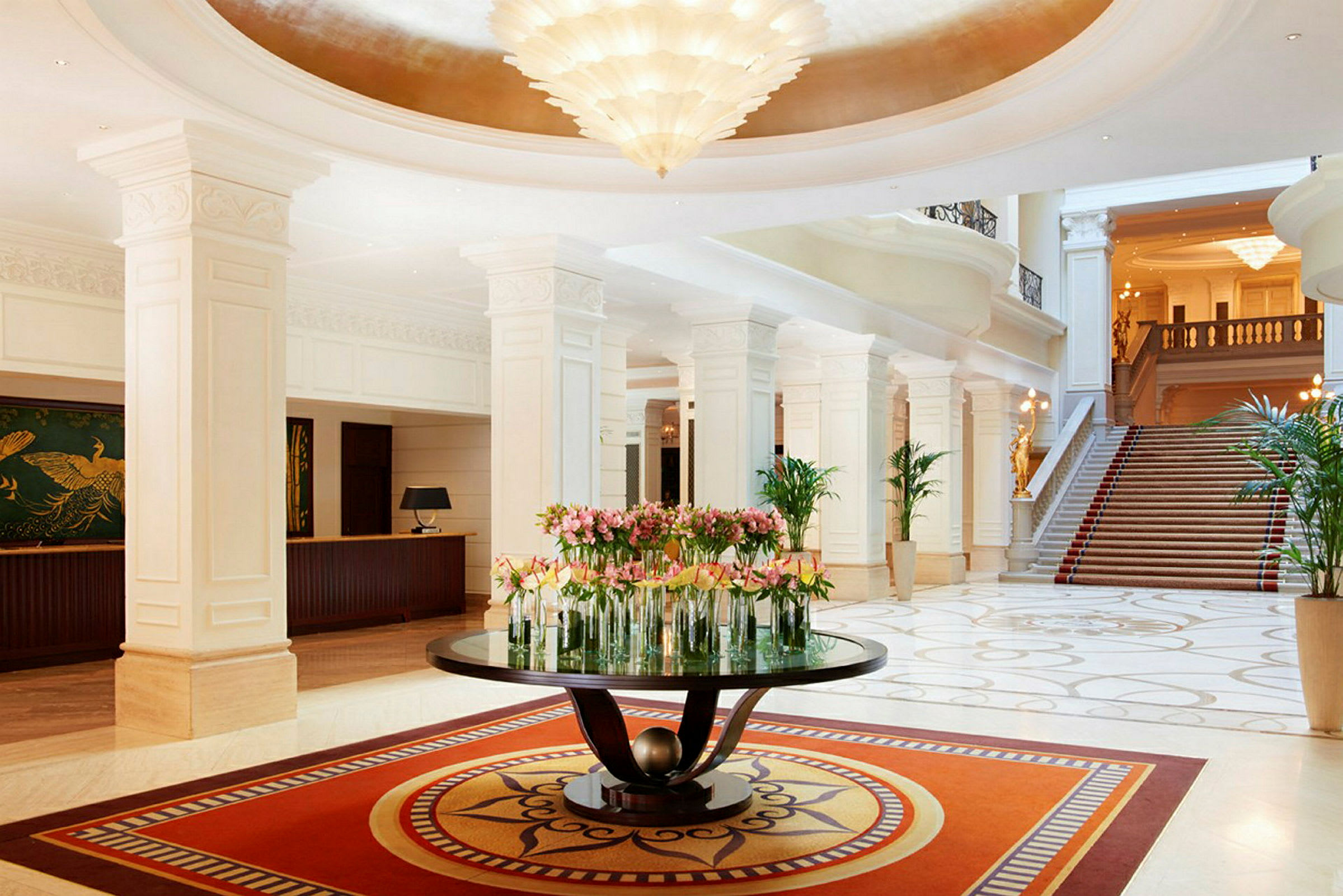 Et elegant hotellobby med marmorgulv, et bord i midten dekorert med blomster og en trapp i bakgrunnen. (AI-generert tekst)