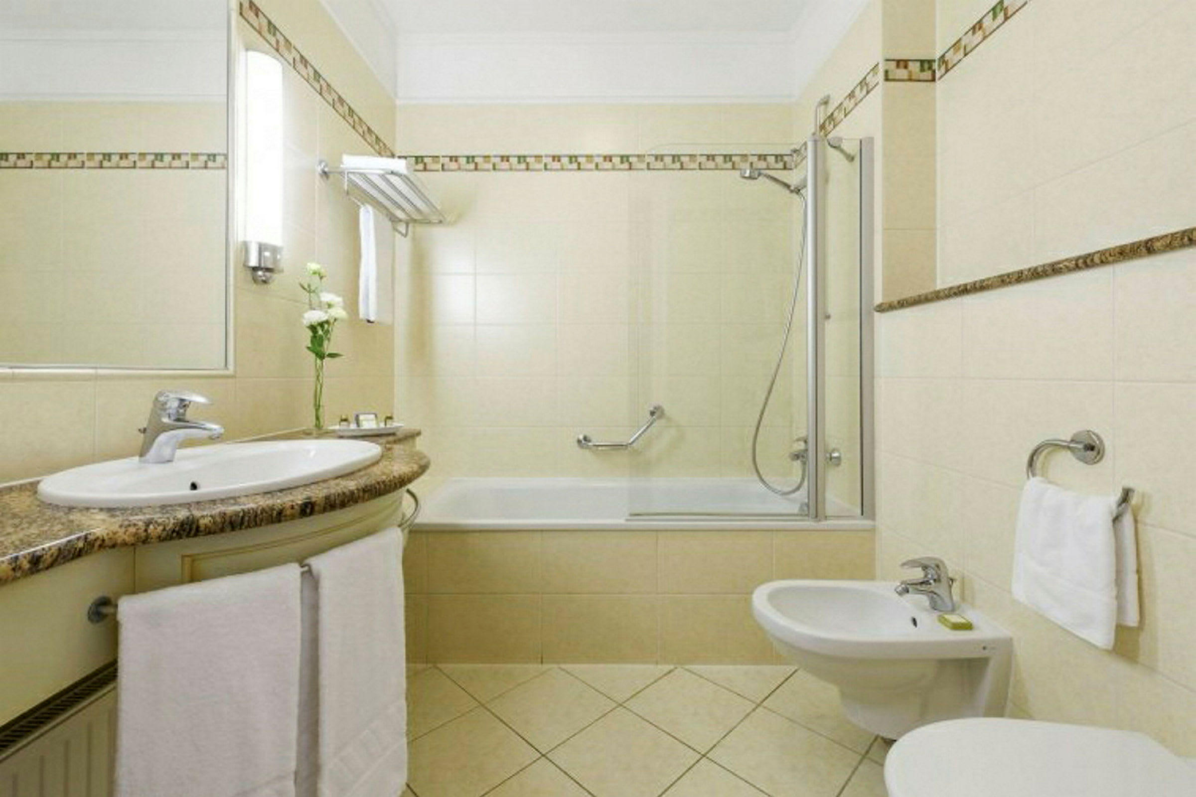 Et baderom med beige fliser, en servant med speil over, et badekar med dusj, en bidet og en håndklestang. (AI-generert tekst)