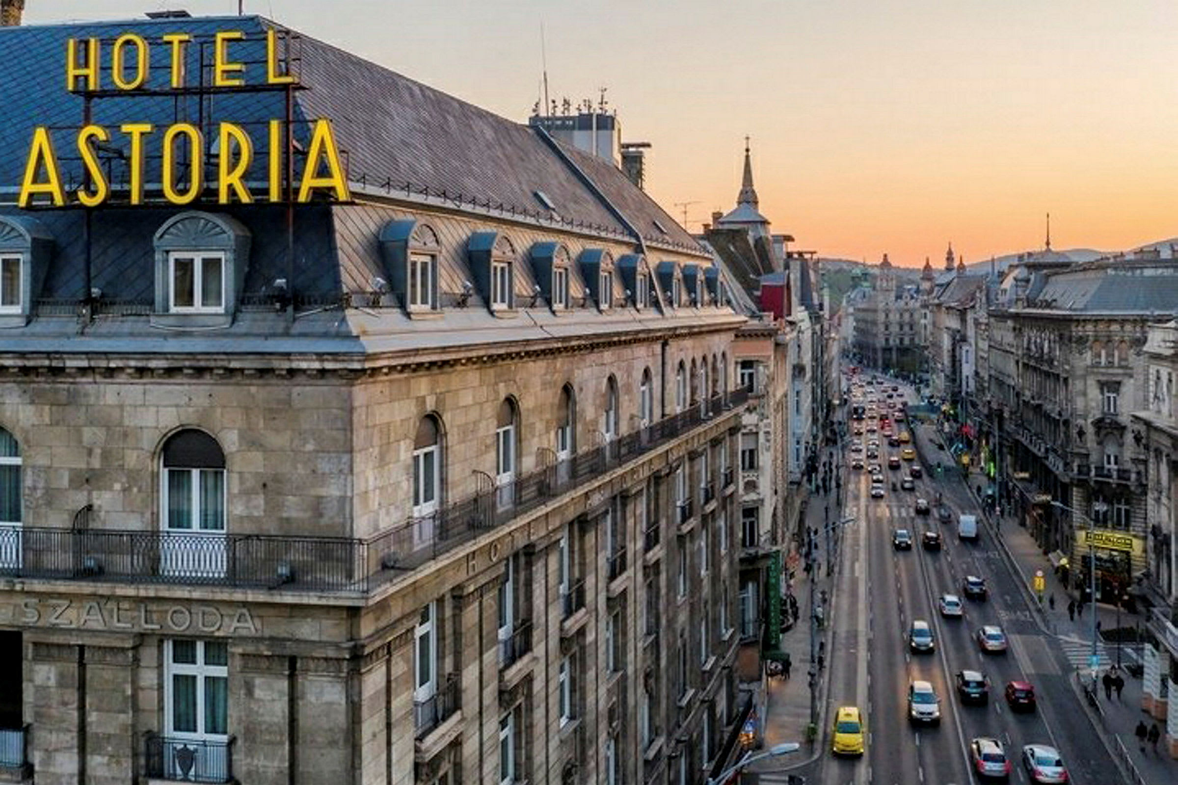 Utsikt over en bygning med teksten "HOTEL ASTORIA" og en travel gate i bymiljø under en solnedgang. (AI-generert tekst)