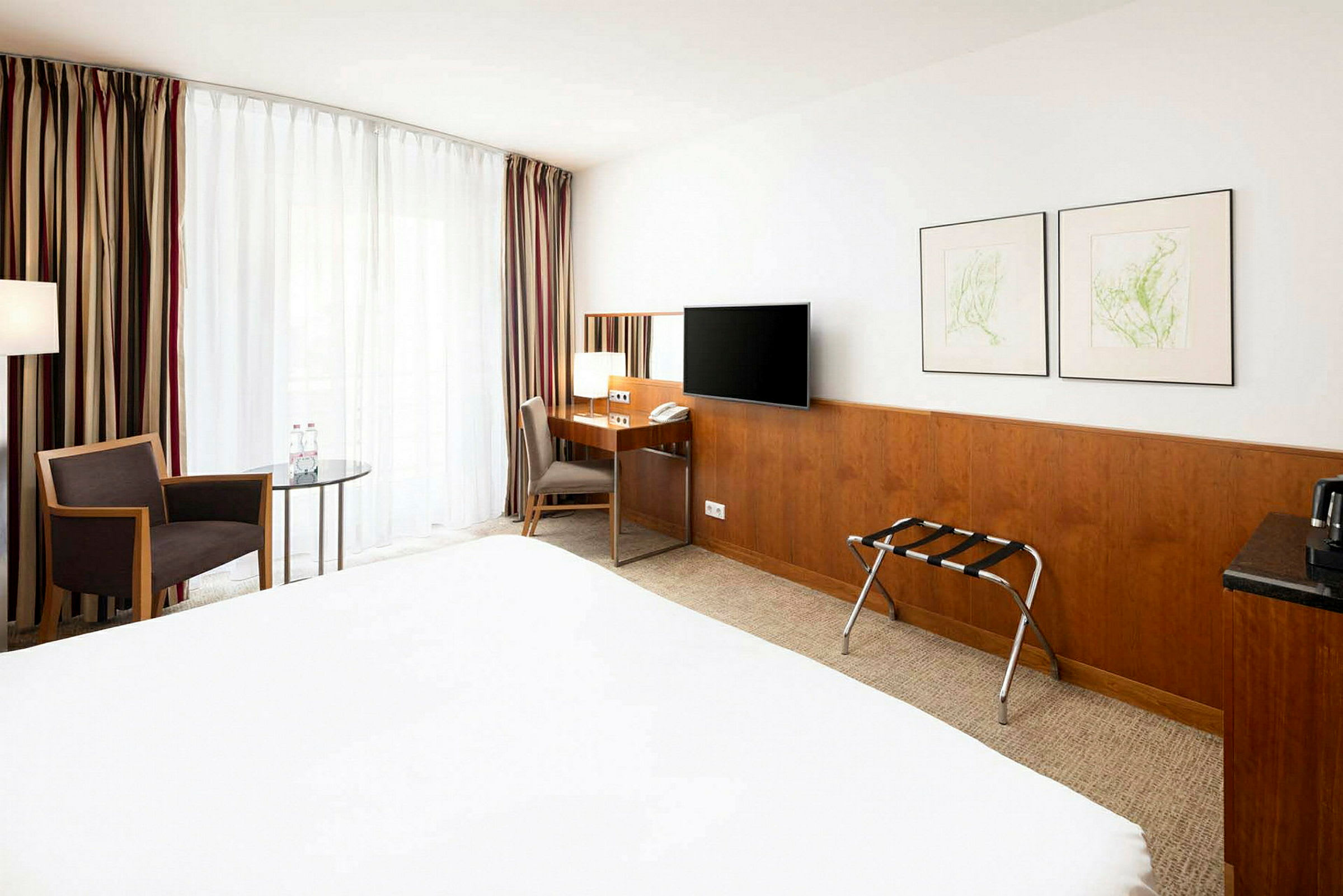 Et hotellrom med skrivebord, stol, TV, to malerier på veggen og en stol ved et bord foran et stort vindu med gardiner. (AI-generert tekst)