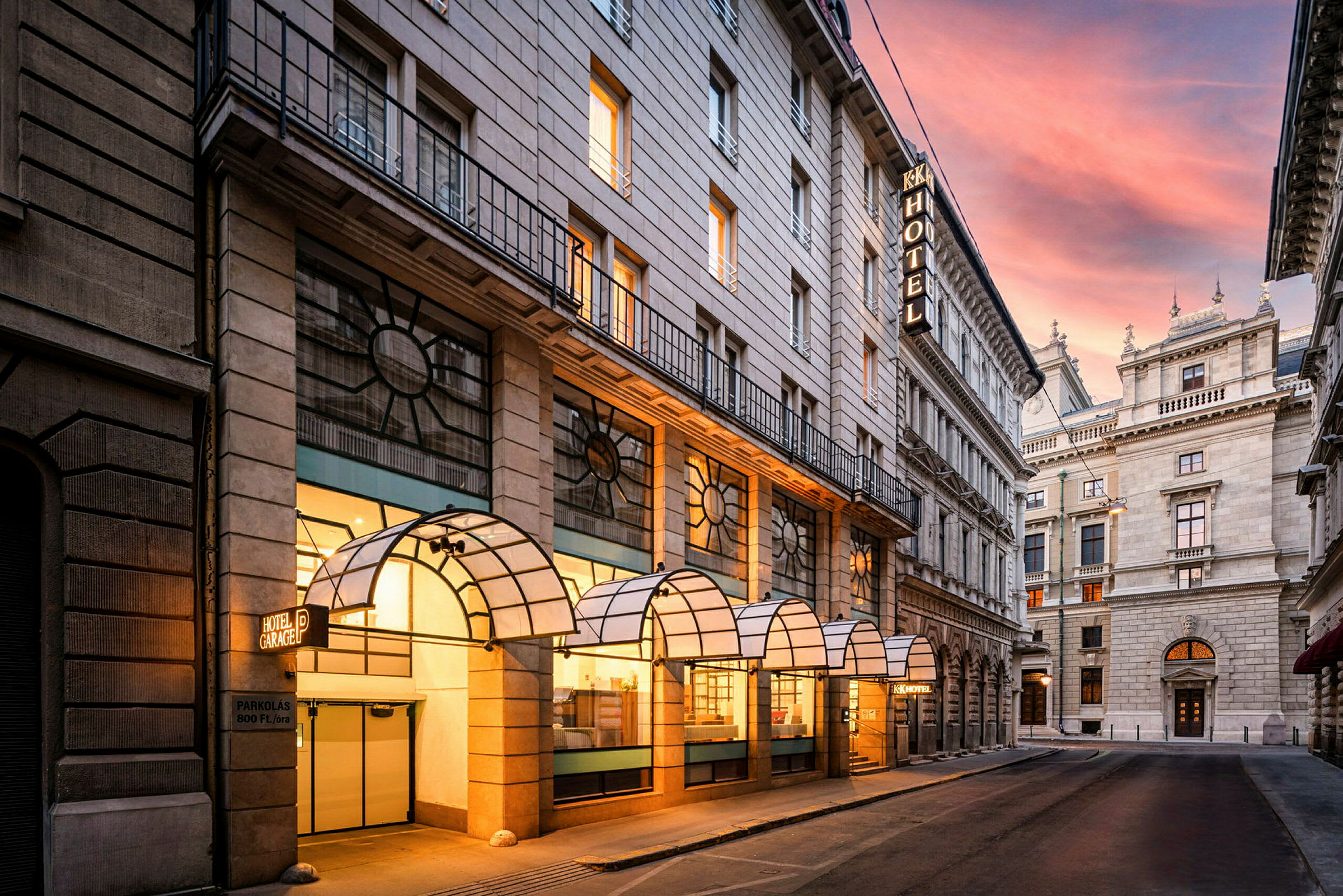 Fasaden til et hotell med overbygde inngangspartier langs en smal bygate. En solnedgang kan ses over gaten. (AI-generert tekst)