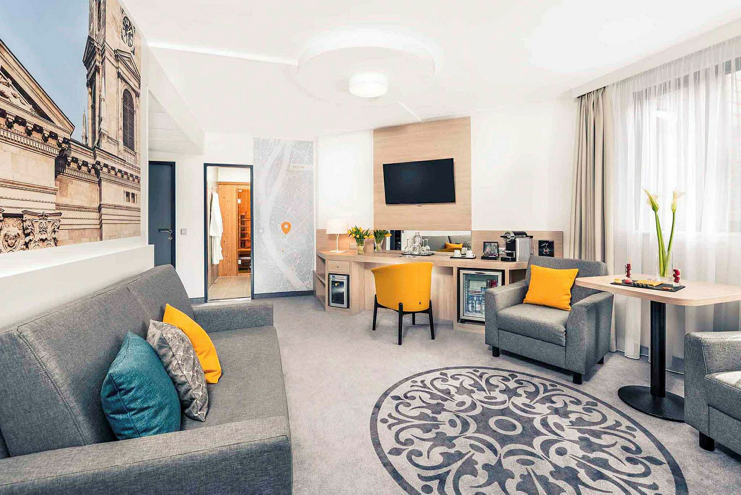Et hotellrom med sittegruppe, skrivebord og et stort veggbilde av en bygningsfasade. (AI-generert tekst)