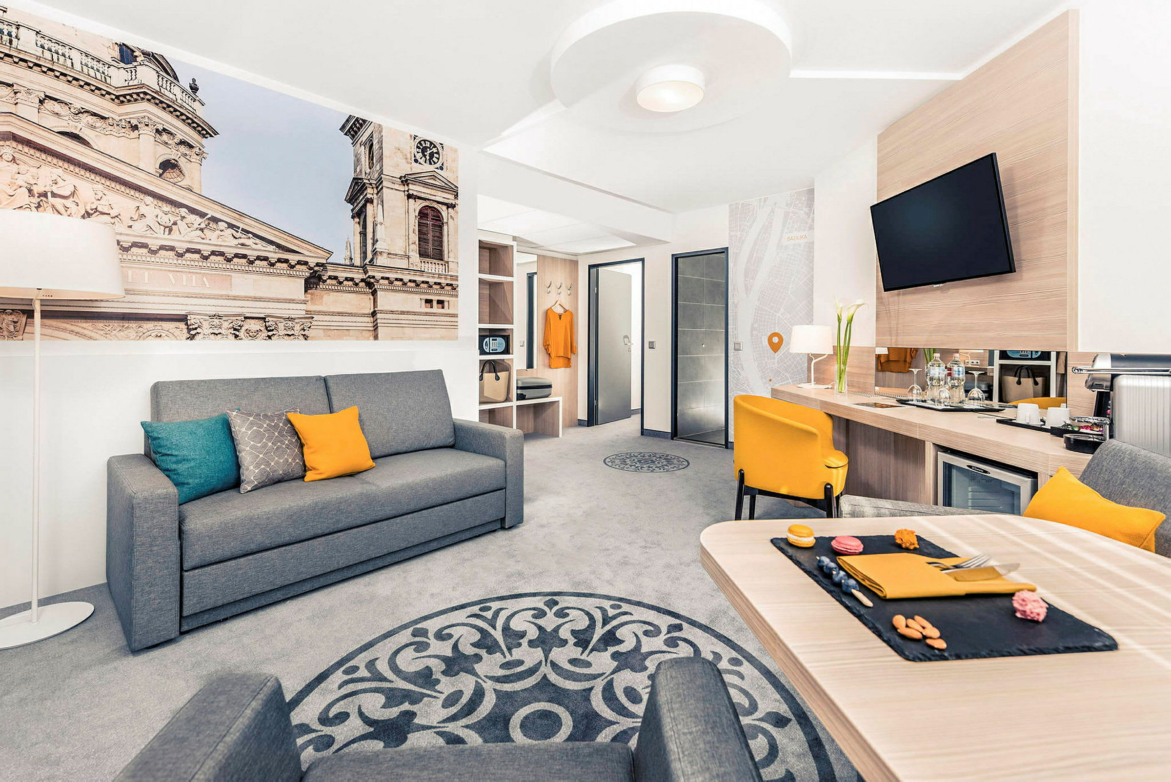 Interiør av et moderne hotellrom med en grå sofa, gul stol og et bord, samt et stort veggbilde som viser en bygning. (AI-generert tekst)