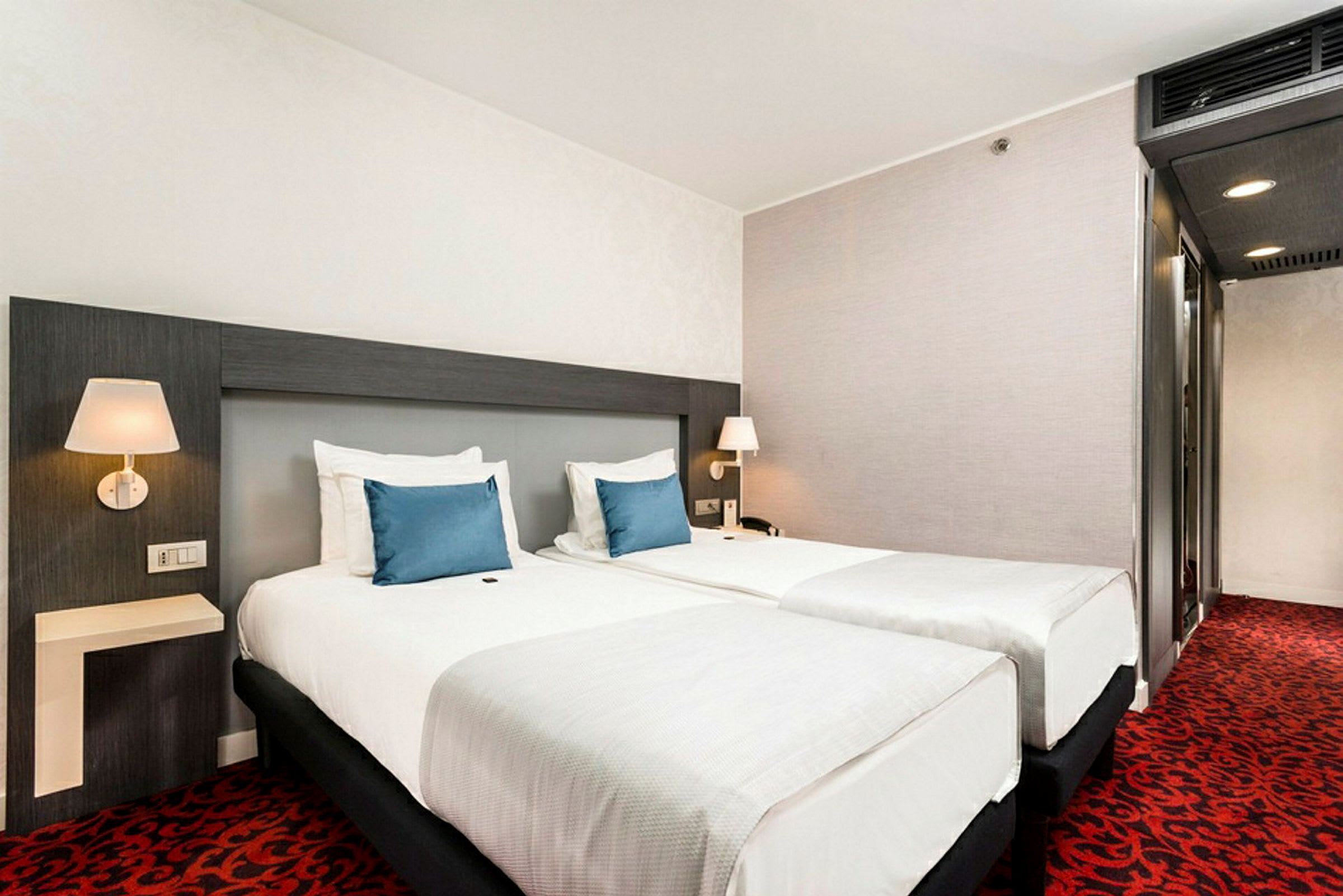 Hotellrom med to separate senger og blå pynteputer. (AI-generert tekst)