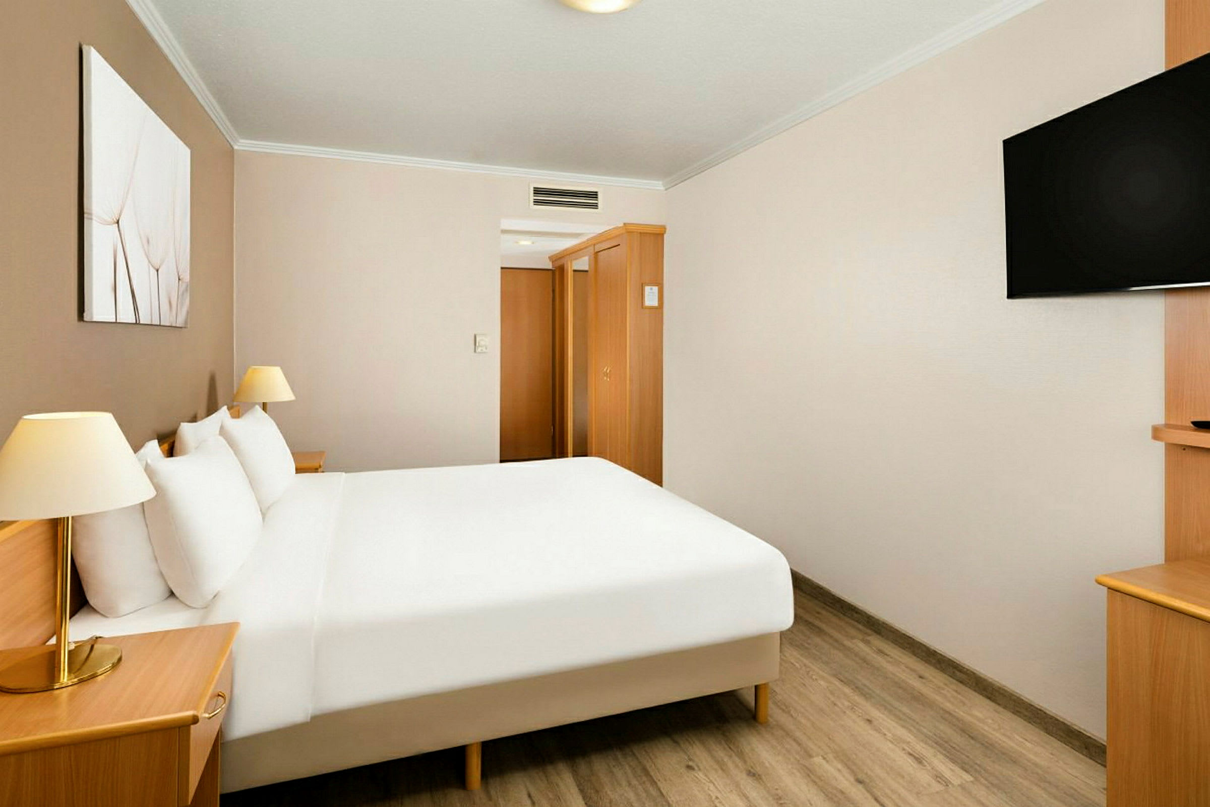 Et hotellrom med en dobbeltseng, hvite laken, nattbord med lamper, veggmontert TV og tremøbler. (AI-generert tekst)