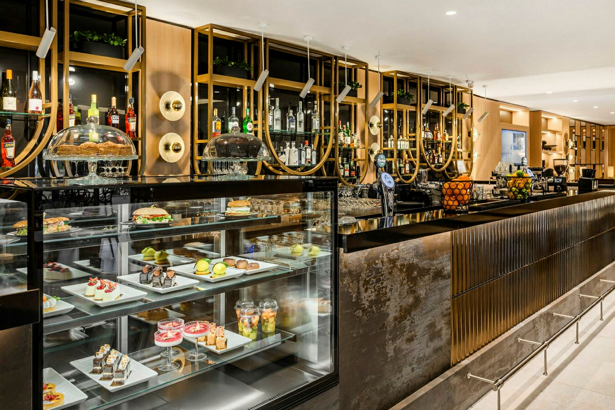 En elegant bardisk med innebygde glassdisplayer som viser forskjellige desserter og frukt. Bak baren kan man se hyller med spritflasker og dekorative elementer. (AI-generert tekst)