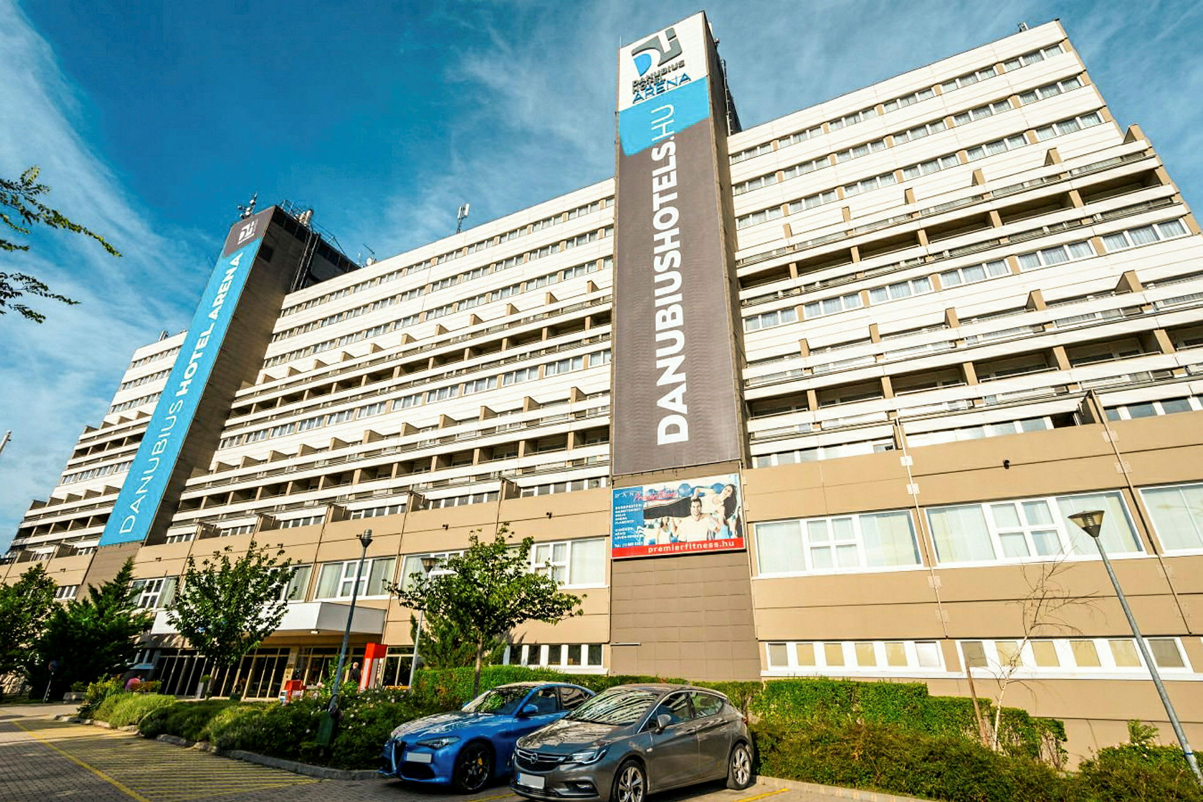En stor hotellbygning med flere etasjer med skiltet ”Danubius Hotel Arena” synlig. Foran bygningen er parkerte biler og noen små trær. (AI-generert tekst)