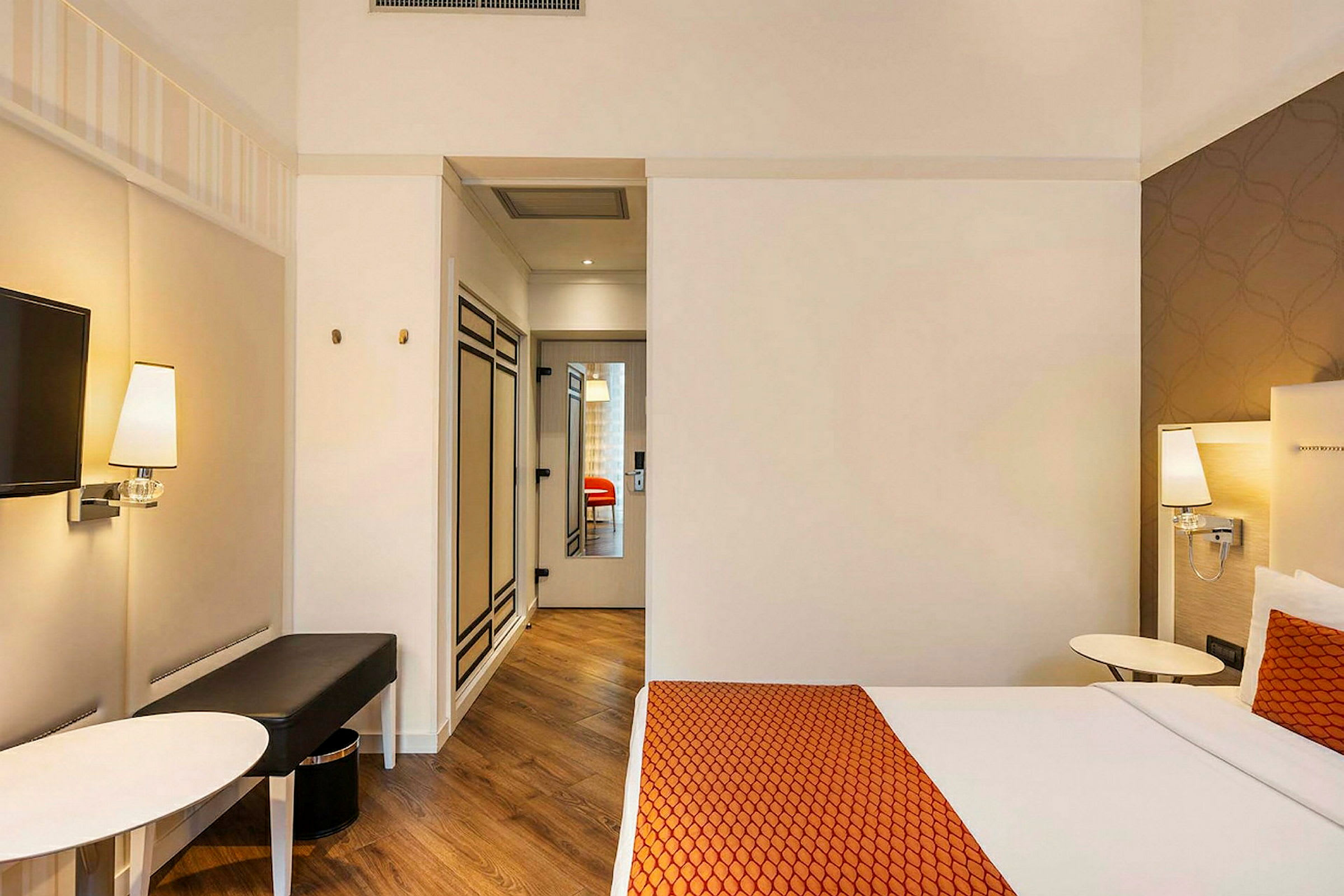 Et hotellrom med en seng med oransje sengeteppe og pute, veggmontert lampe, bord og TV, samt en inngang med speil og tregulv. (AI-generert tekst)