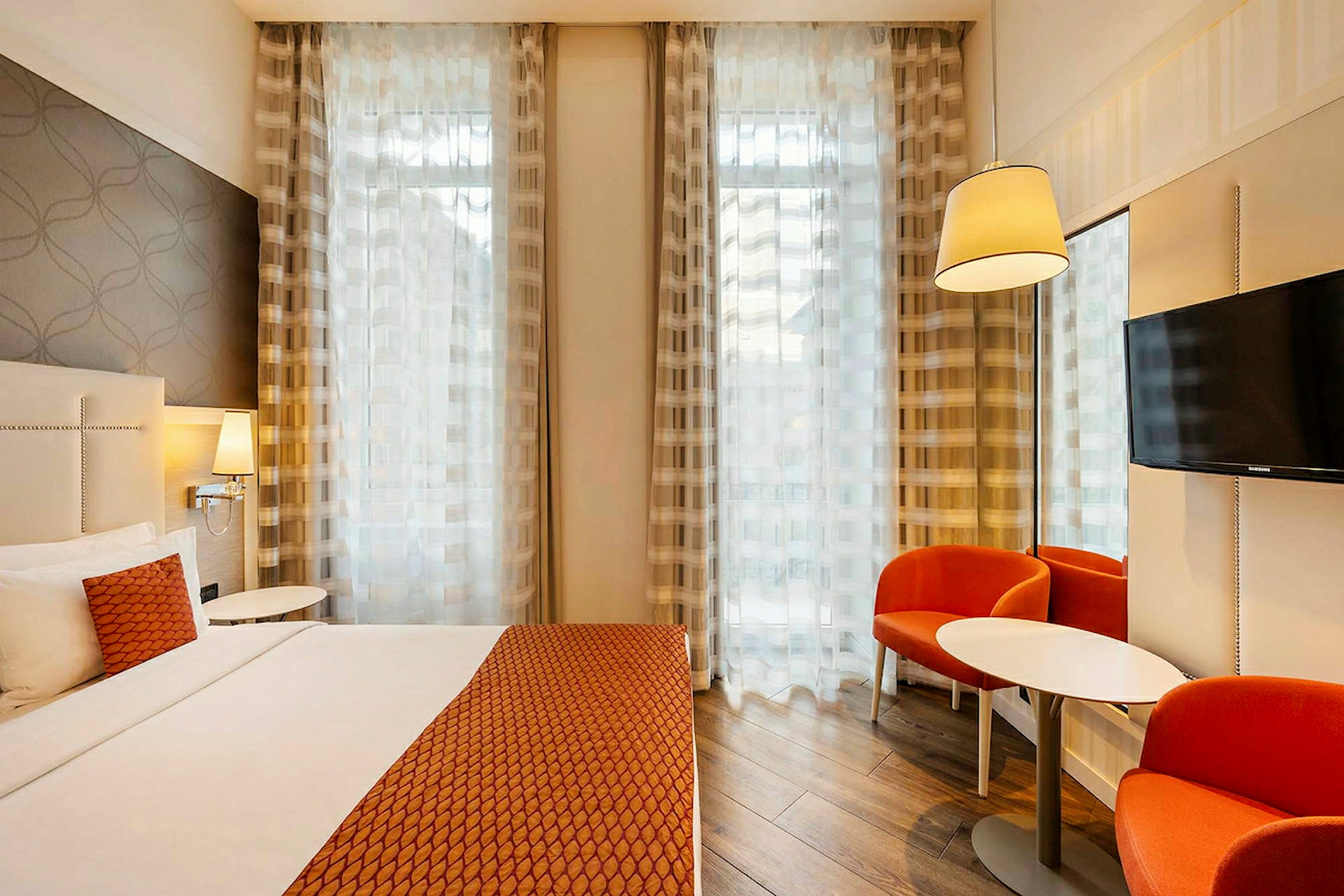 Hotell soverom med dobbeltseng med oransje sengeteppe og puter. To panelgardiner dekker store vinduer, en bordlampe og en veggmontert TV. To oransje stoler og et rundt bord står i hjørnet. (AI-generert tekst)