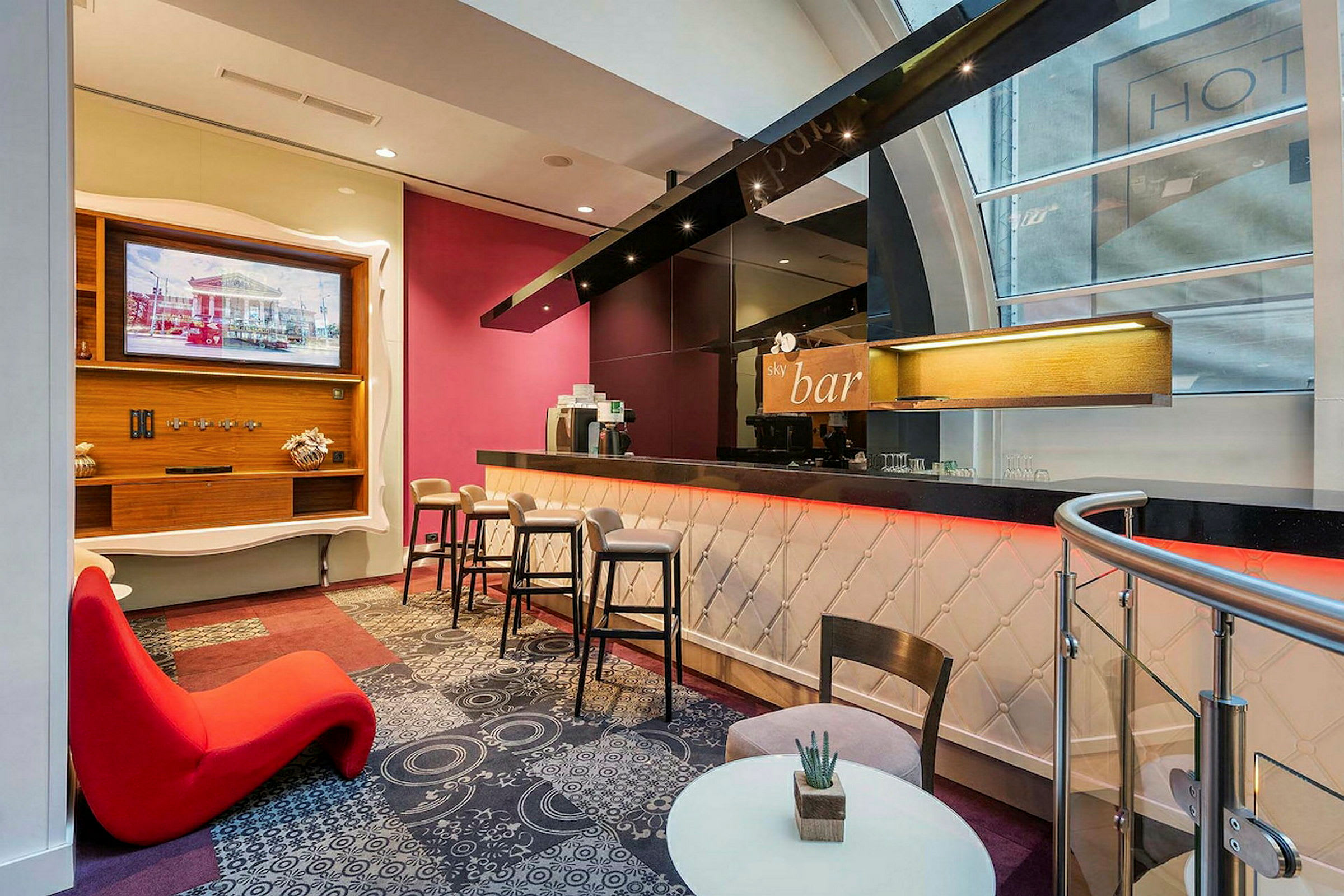 Bildet viser en moderne bar inne i en bygning med stilfull innredning, inkludert bardstoler, lounge-stoler, en overhengende hylleenhet og detaljert teppe. (AI-generert tekst)