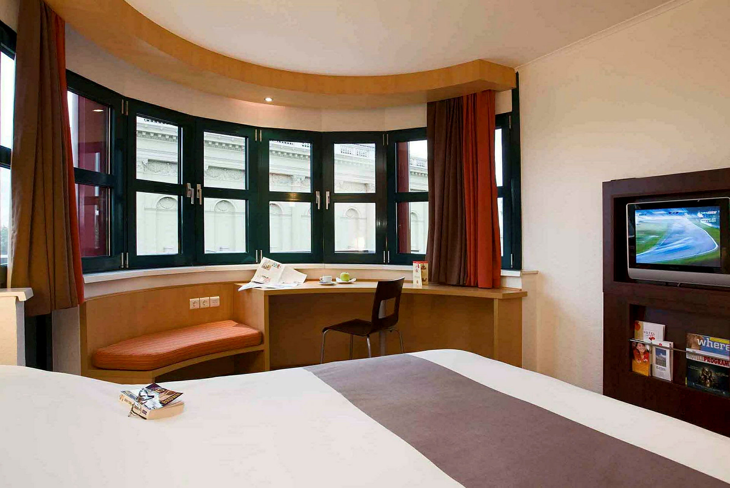 Et moderne hotellrom med en stor seng, et skrivebord foran store vinduer med utsikt mot en bygning og en TV som står på et møbel. (AI-generert tekst)