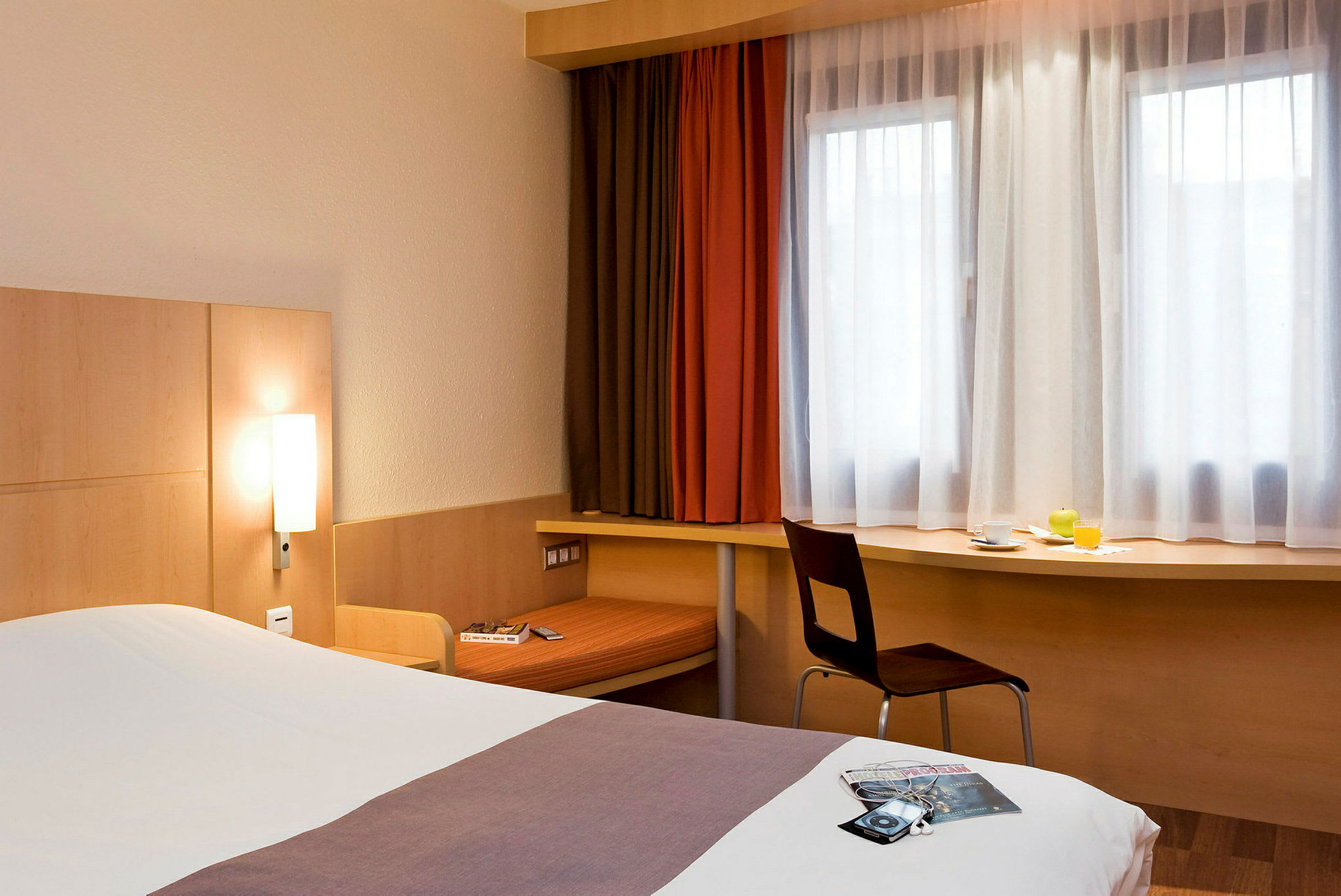Et hotellrom med en seng, et skrivebord under vinduet og en stol. Sengen er oppredd, og det står en kopp, et glass appelsinjuice og en grønn frukt på skrivebordet. (AI-generert tekst)