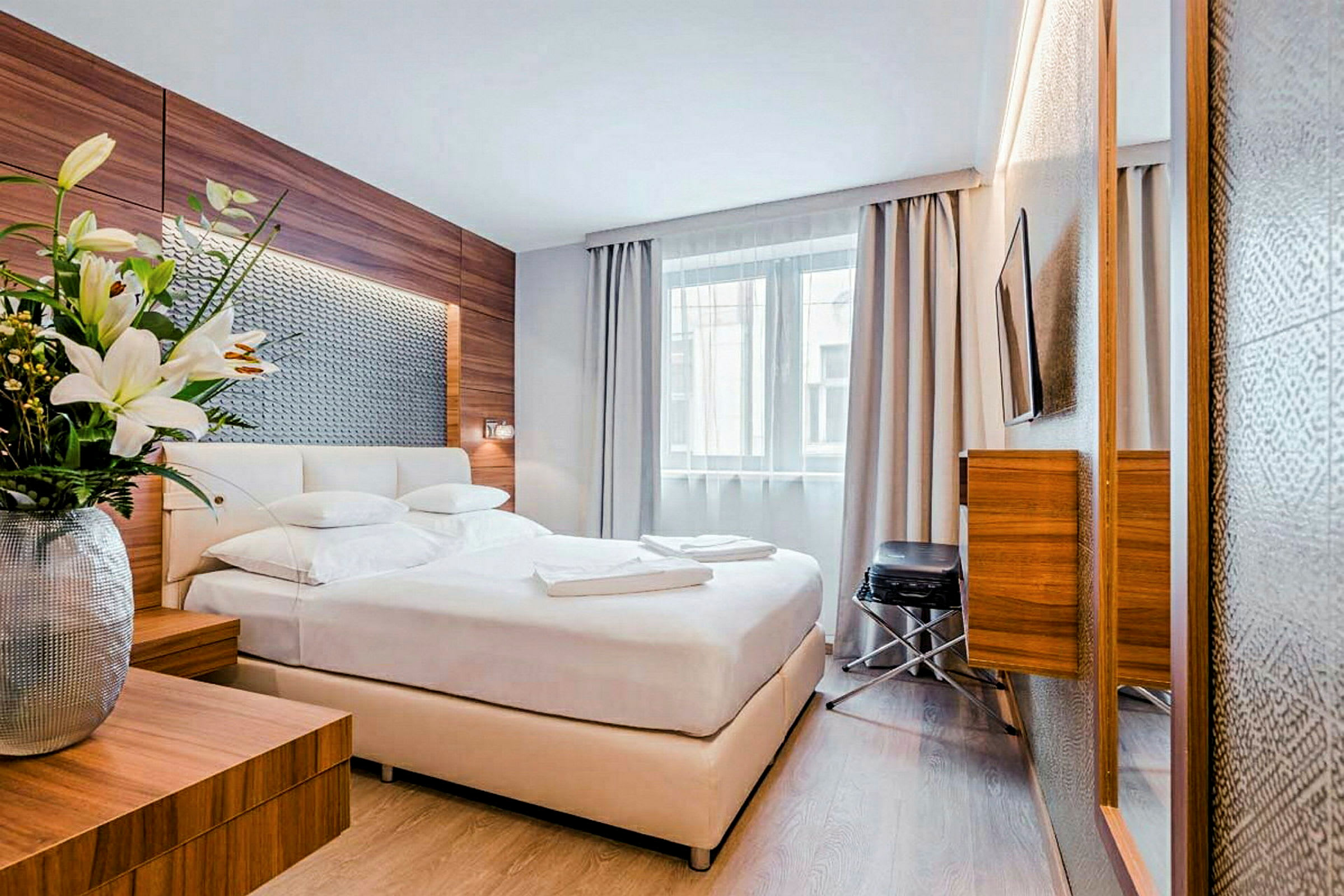 Et moderne hotellrom med en dobbeltseng, et stort vindu med gardiner og frisk innredning med tre detaljer. En vase med blomster står på et nattbord. (AI-generert tekst)