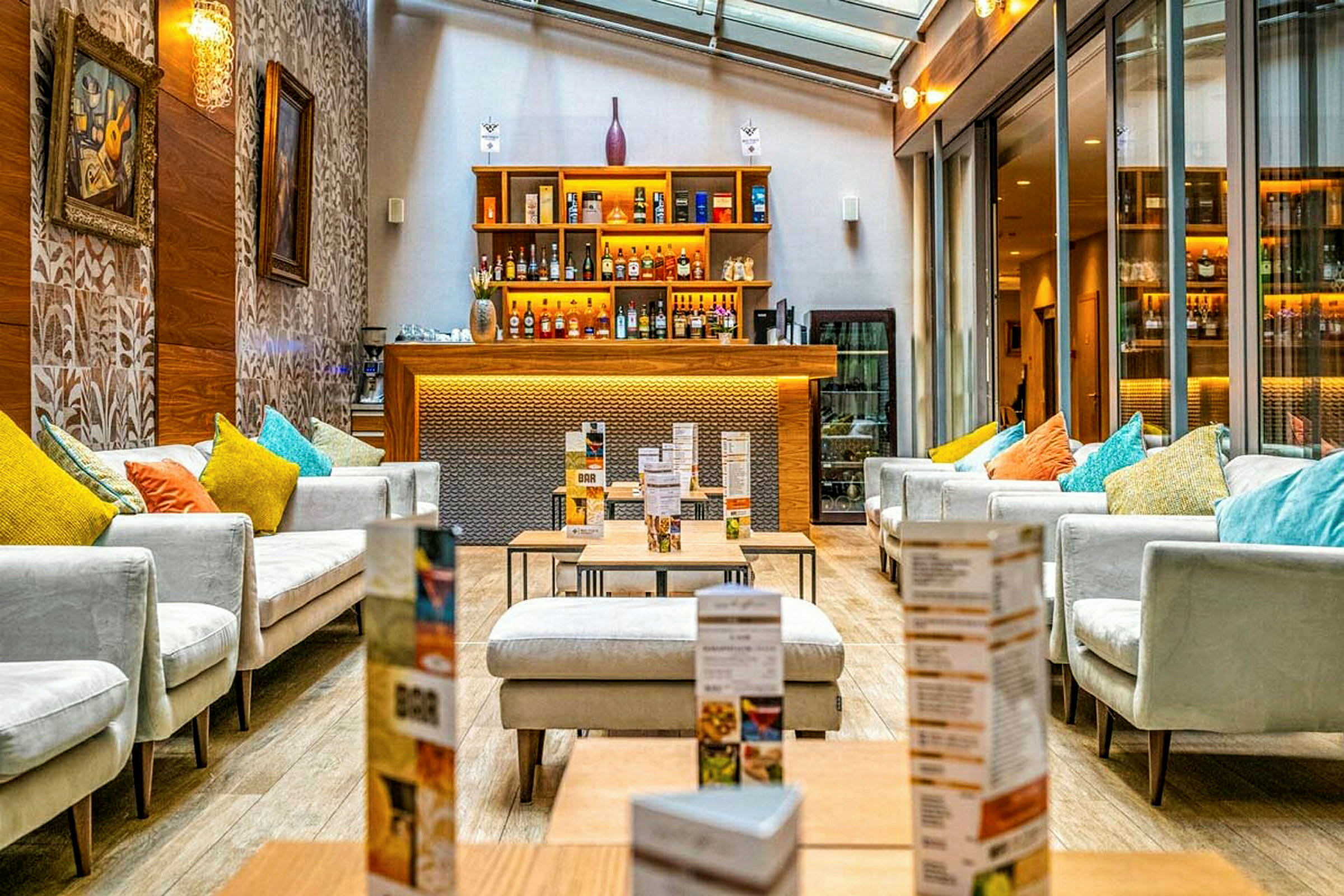 Et moderne loungeområde med sofaer og puter i ulike farger, bord og en bar med opplyste hyller fylt med flasker. (AI-generert tekst)