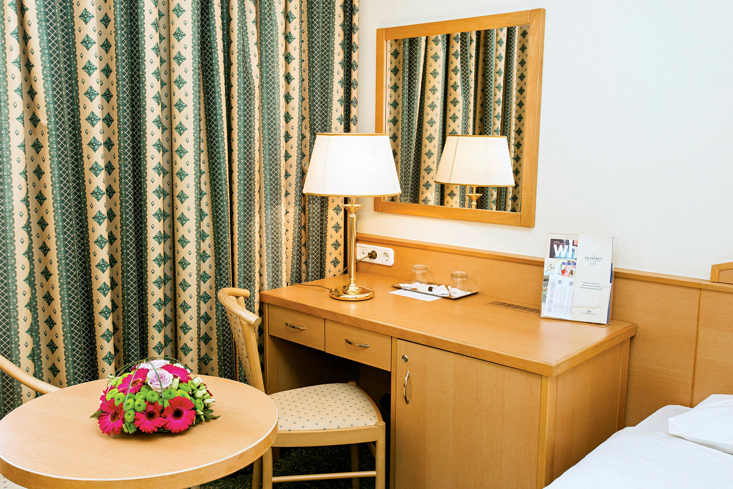 Et hotellrom med et skrivebord, speil, bord med blomsterbukett og lampe. (AI-generert tekst)