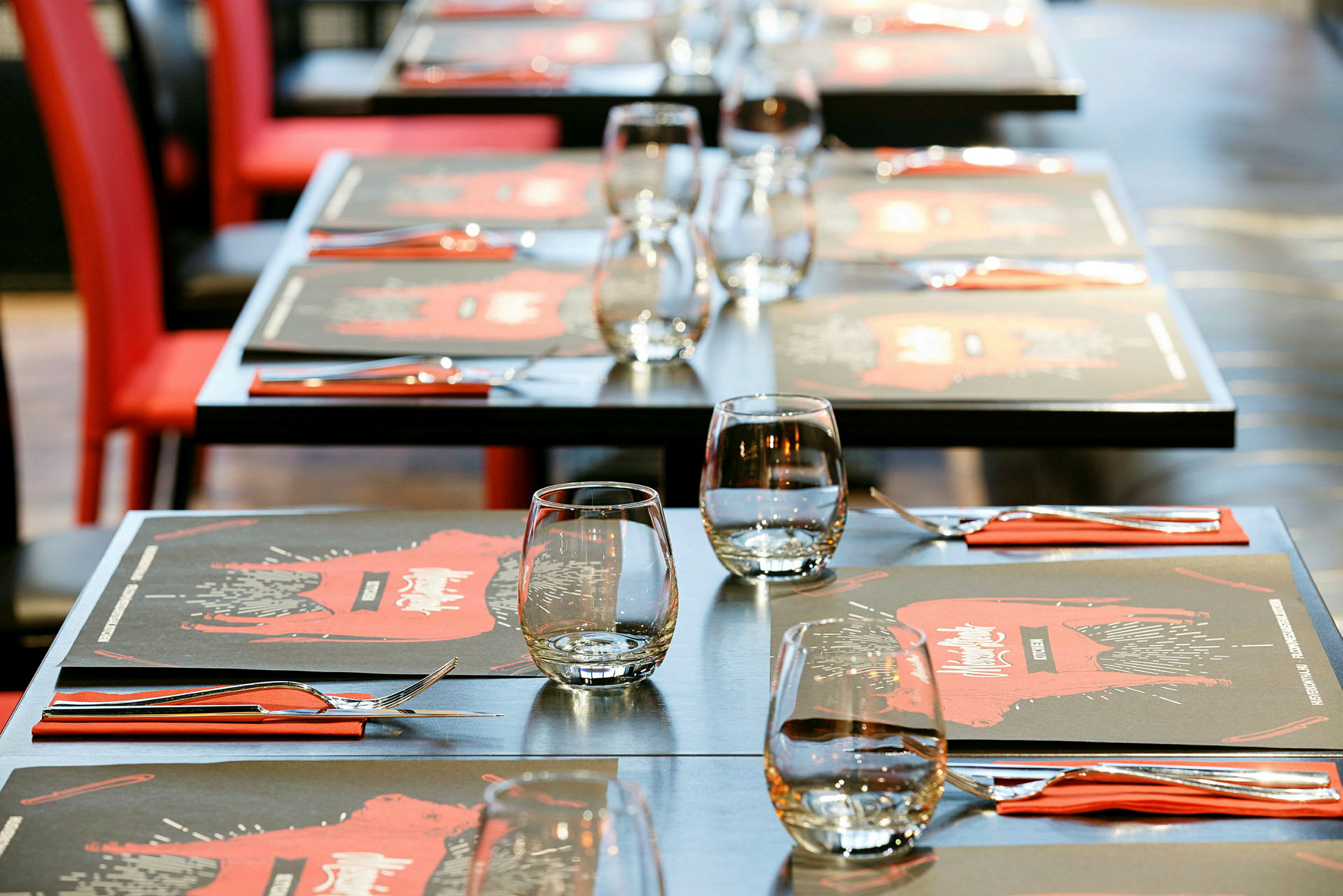 Oppstilte bord med glass, bestikk og røde servietter, restaurantmiljø. (AI-generert tekst)